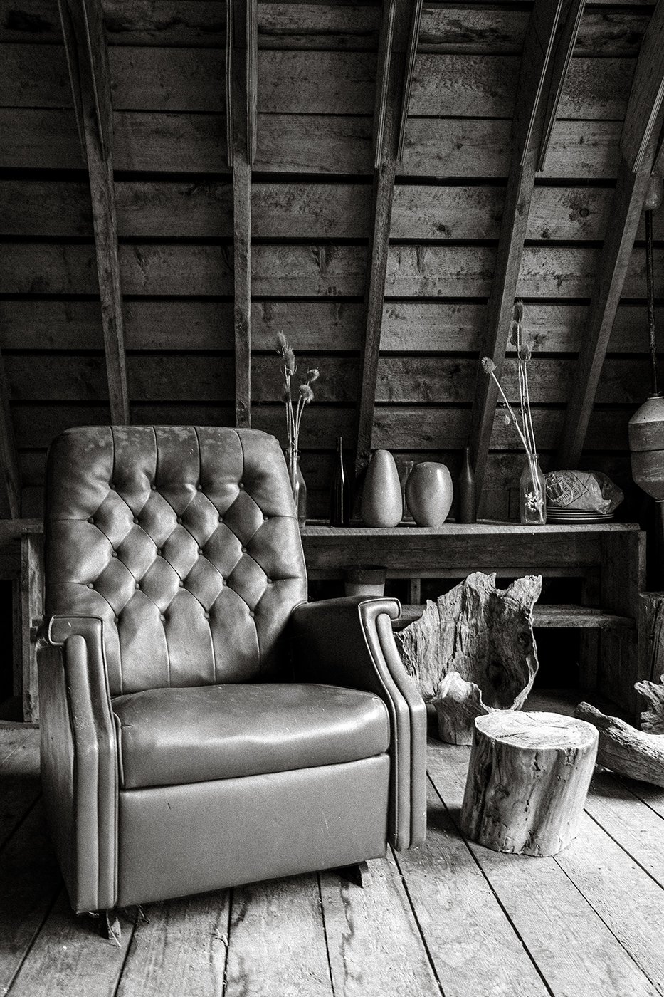 A vintage leather armchair in an attic with wooden walls and floor, surrounded by natural wood logs, pottery vases, and dried plants in vases on a shelf.