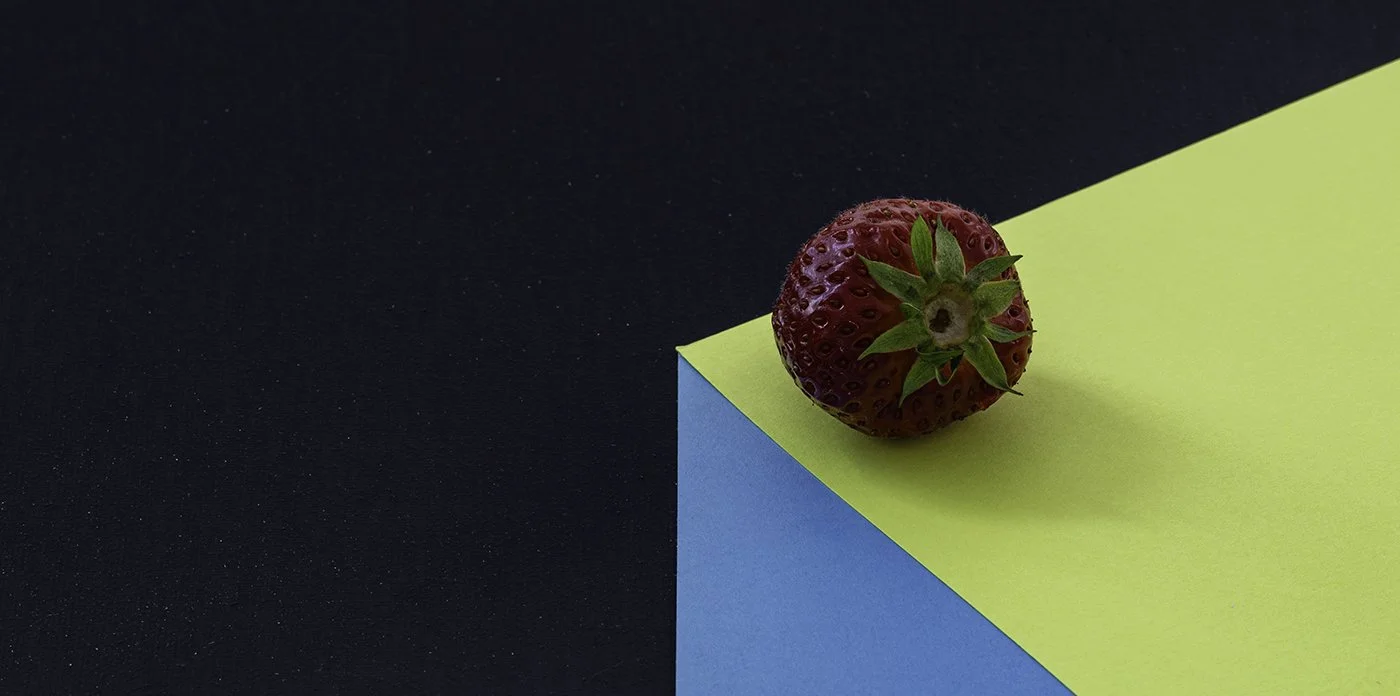 A ripe red strawberry with green leaves resting on a corner of yellow and blue paper on a black surface.