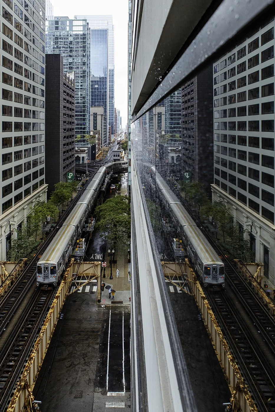 A city street with high-rise buildings, an elevated train track, and a moving train, seen from a tall building window during rainy weather.