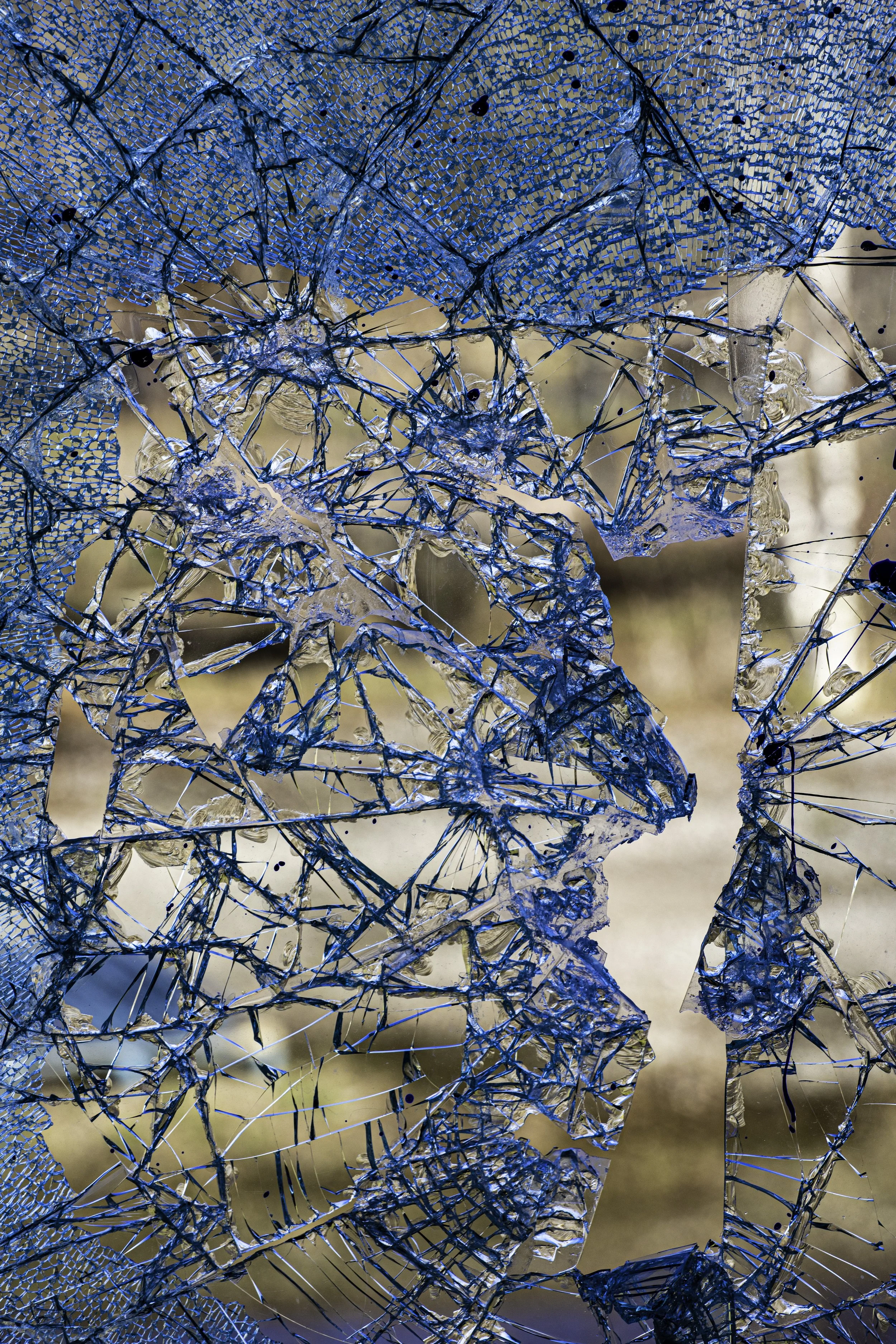 Close-up of shattered blue glass with intricate cracks and broken pieces, with a blurred outdoor background.
