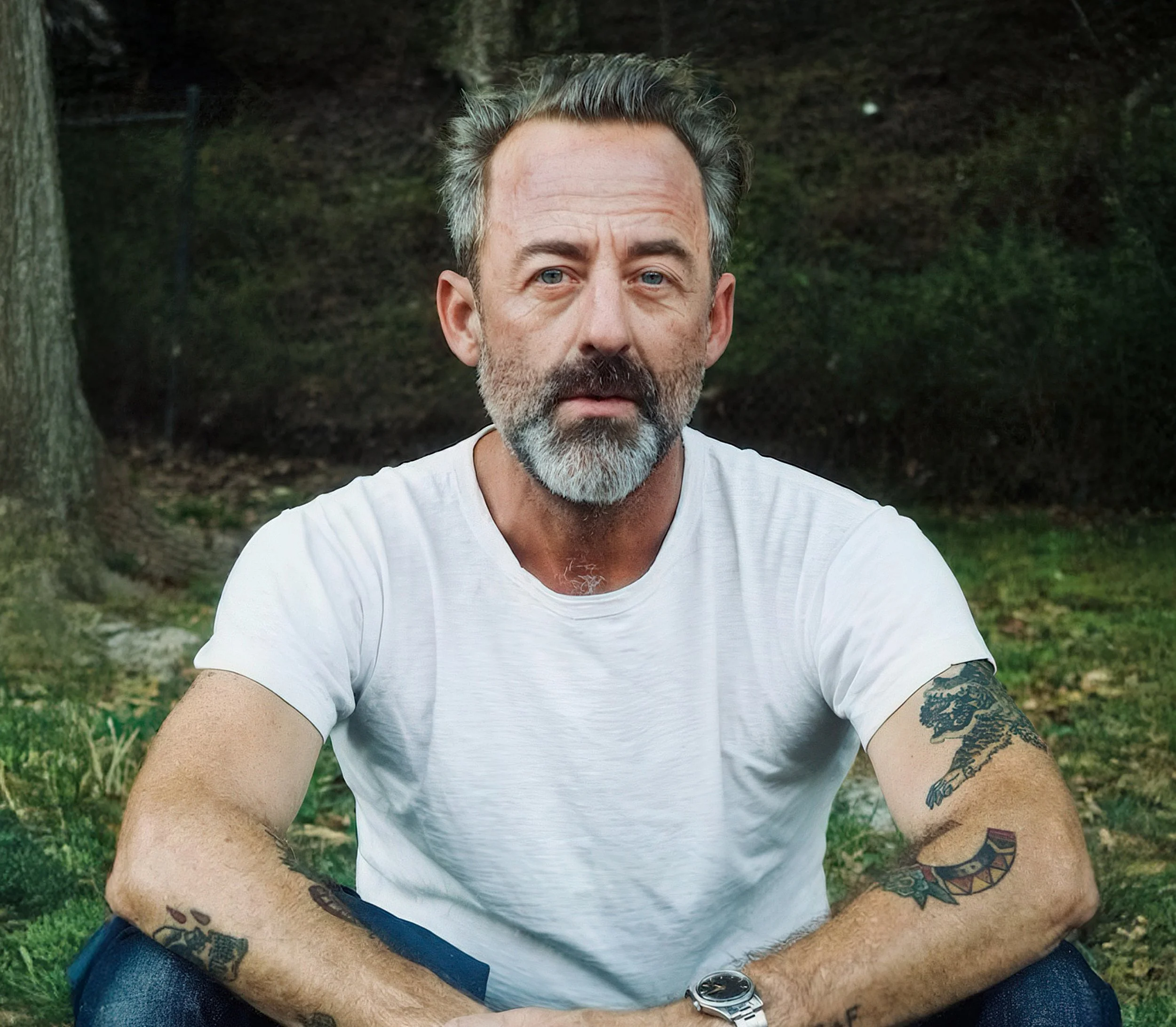 A middle-aged man with gray hair and a beard, wearing a white t-shirt, sitting outdoors on grass with trees in the background.