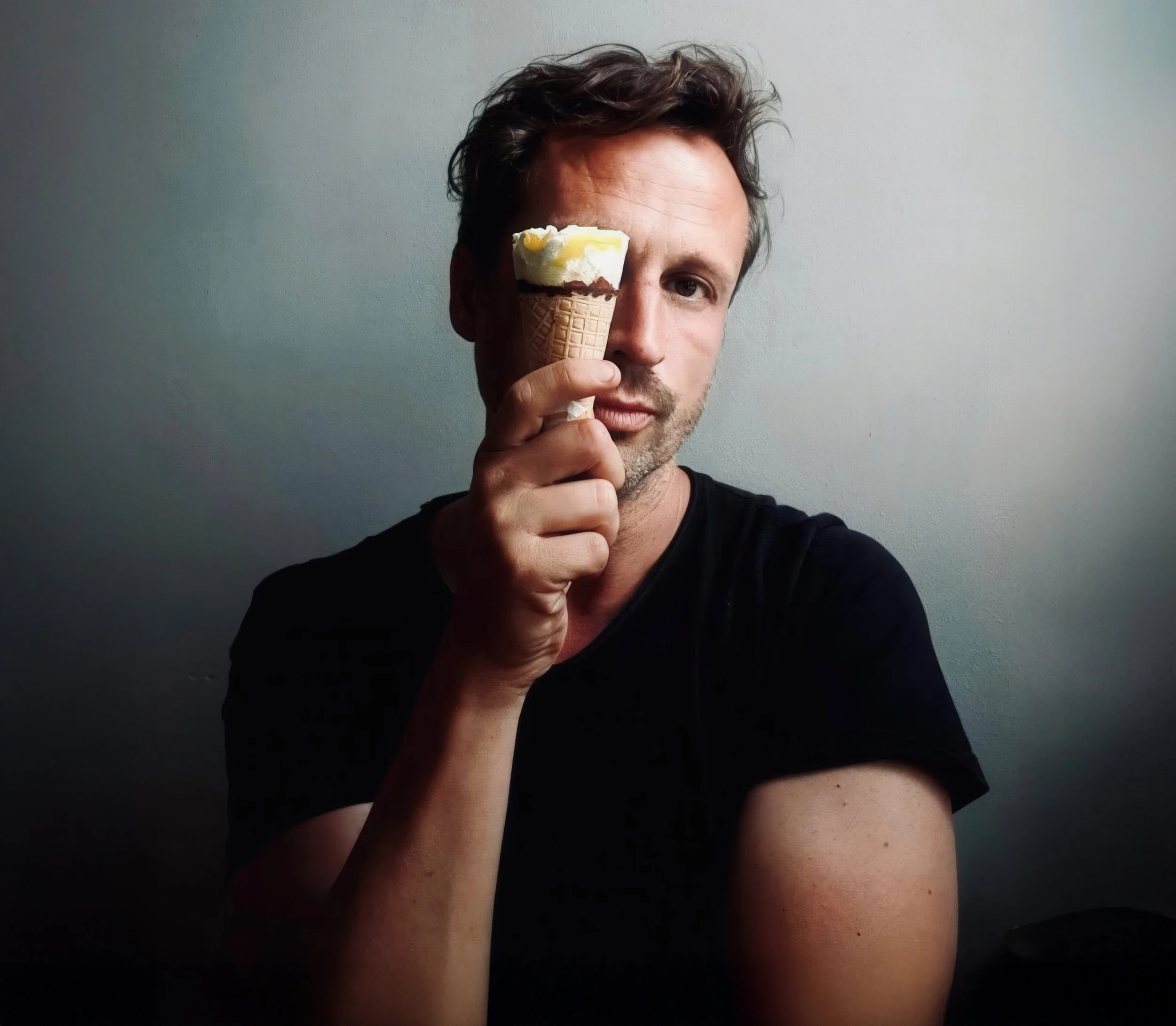 A man with brown hair and a beard holds an ice cream cone in front of his face, covering one eye, against a plain gray background.