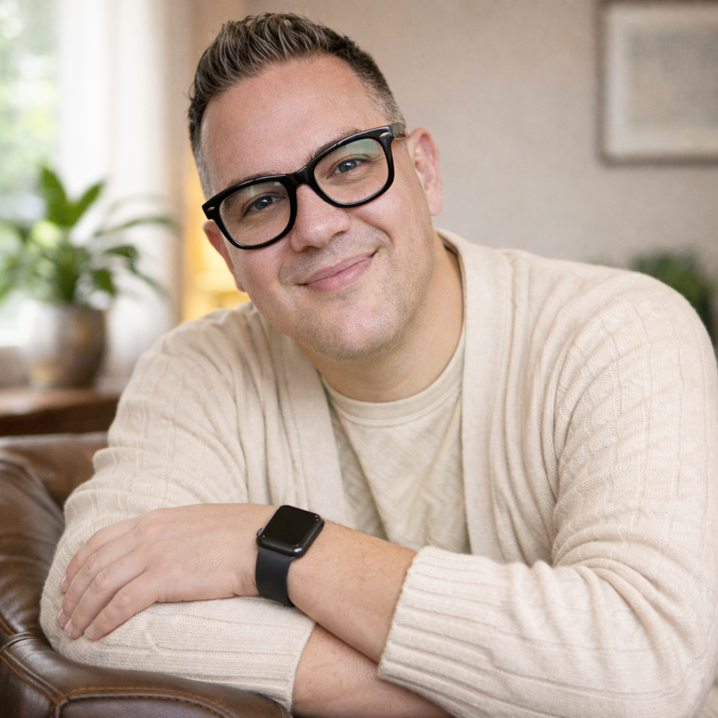 A smiling man wearing glasses and a beige sweater, sitting with his arms crossed on a leather sofa, indoors with a plant and a lamp in the background.