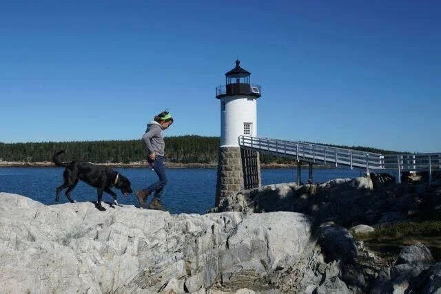 Child with dog exploring the rocky shoreline near Robinson Point lighthouse on Isle au Haut under a clear blue sky.