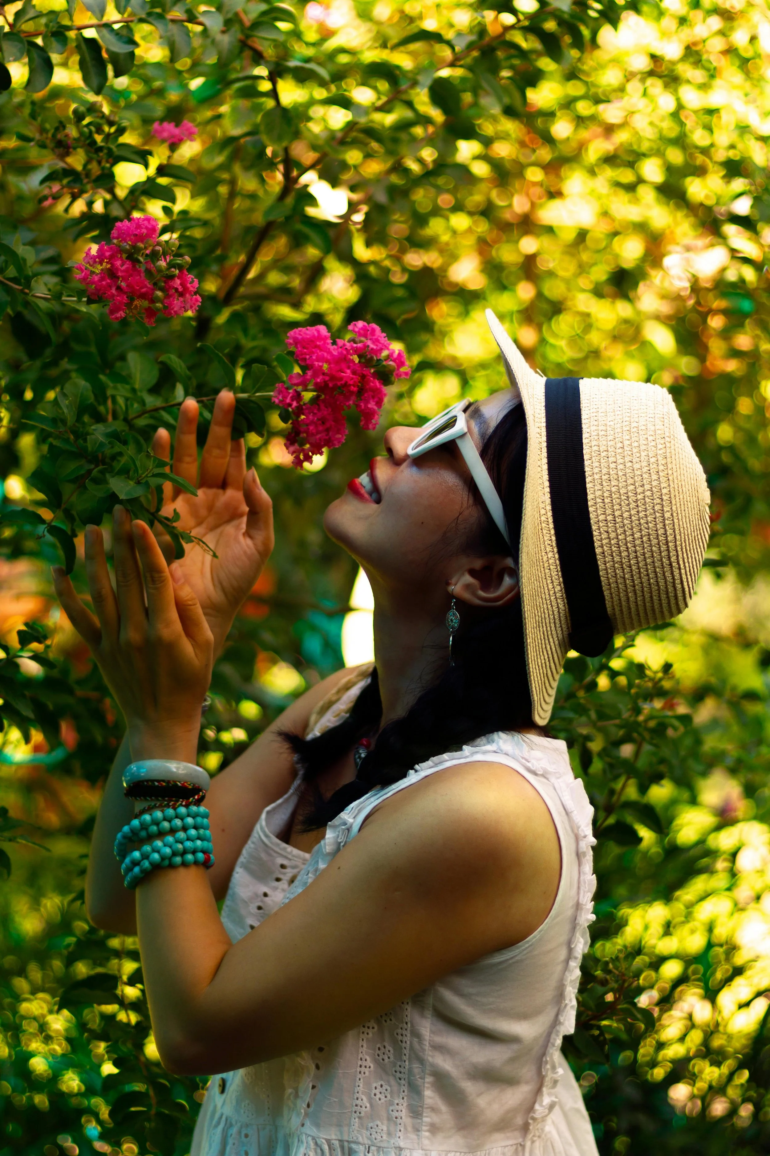 A woman wearing a straw hat, sunglasses, and jewelry, standing outdoors among green foliage and pink flowers, smiling and touching a branch.