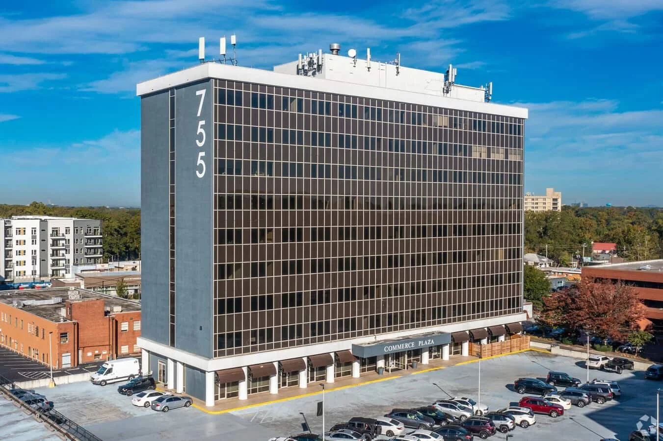 Tall office building with the address 755 visible on the side and a sign reading 'Commerce Plaza' at the entrance, surrounded by a parking lot with several cars, under a blue sky.