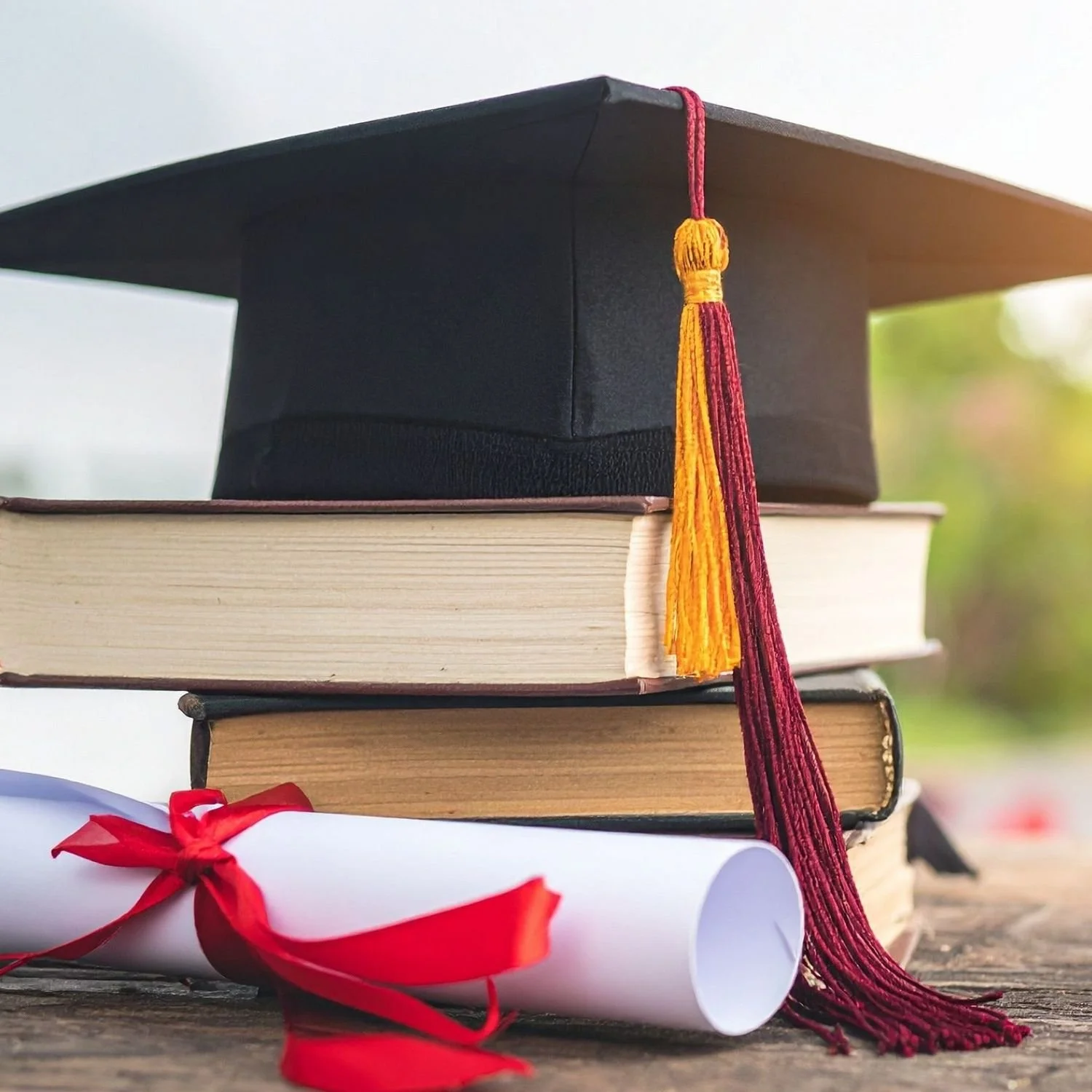 Stack of three books with a graduation cap on top, a diploma scroll with a red ribbon in front, and a blurred background of trees and sky.