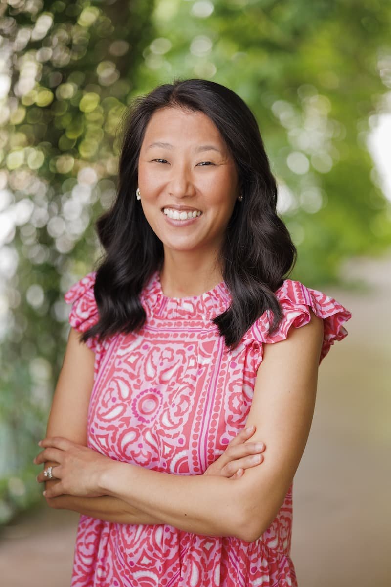 A woman with long black hair smiling and standing outdoors, wearing a pink patterned dress with ruffled sleeves, with green foliage in the background.