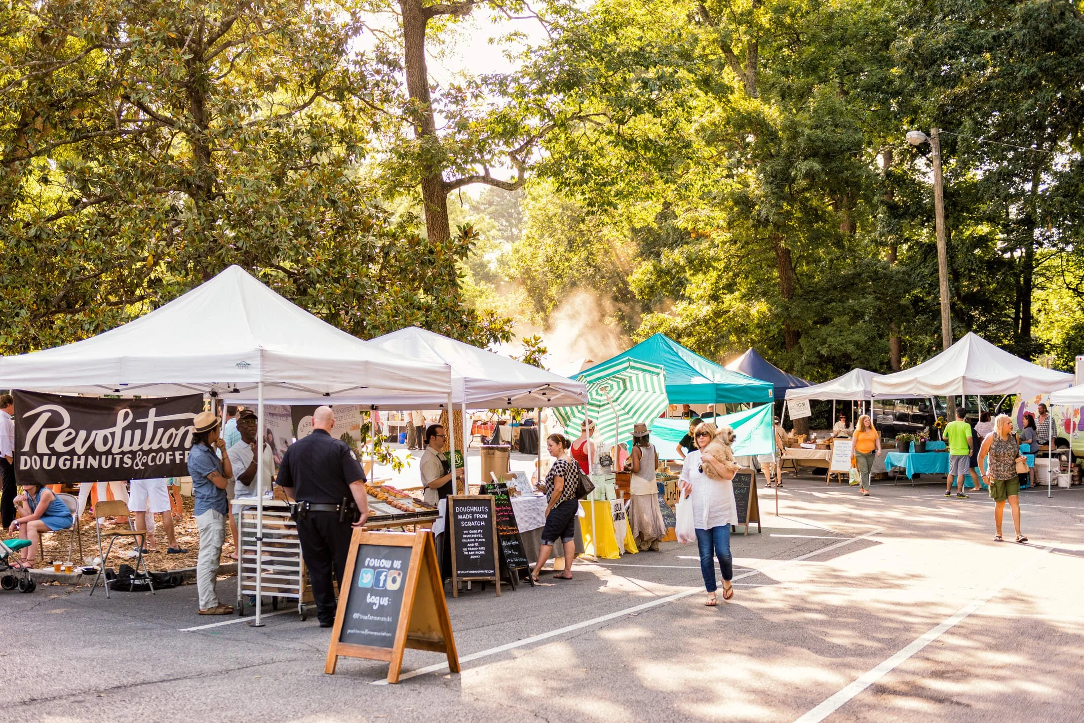 Outdoor market with white tents and people shopping among trees on a sunny day.