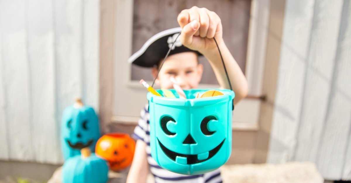 Child in striped shirt holding up a blue pumpkin-shaped Halloween bucket filled with candy.