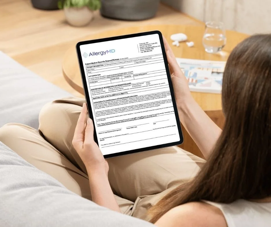 A woman holding a tablet displaying a medical record form from AllergyMD, sitting on a couch in a living room with a wooden table, water glass, and magazines nearby.