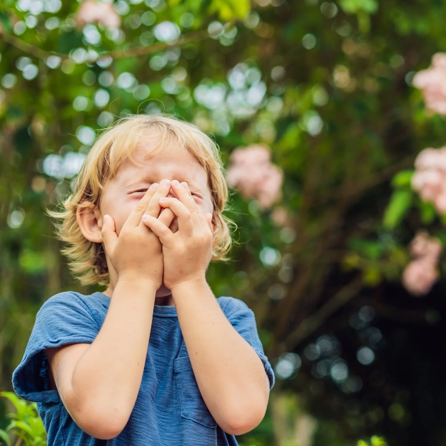 A young boy with blonde hair covering his face with his hands, standing outdoors with green foliage and pink flowers in the background.
