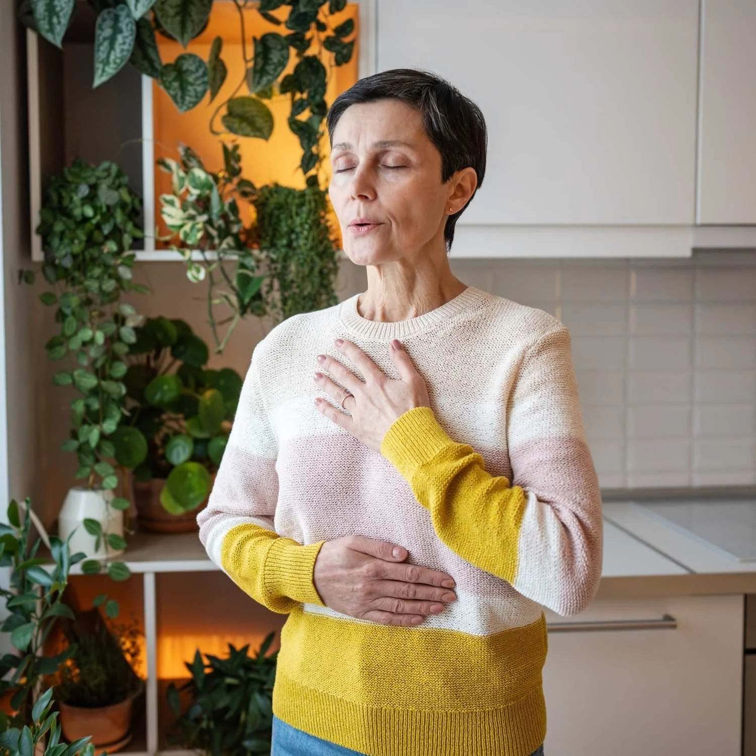A woman with short dark hair holding her chest and stomach, standing in a room with green plants and white kitchen cabinets.