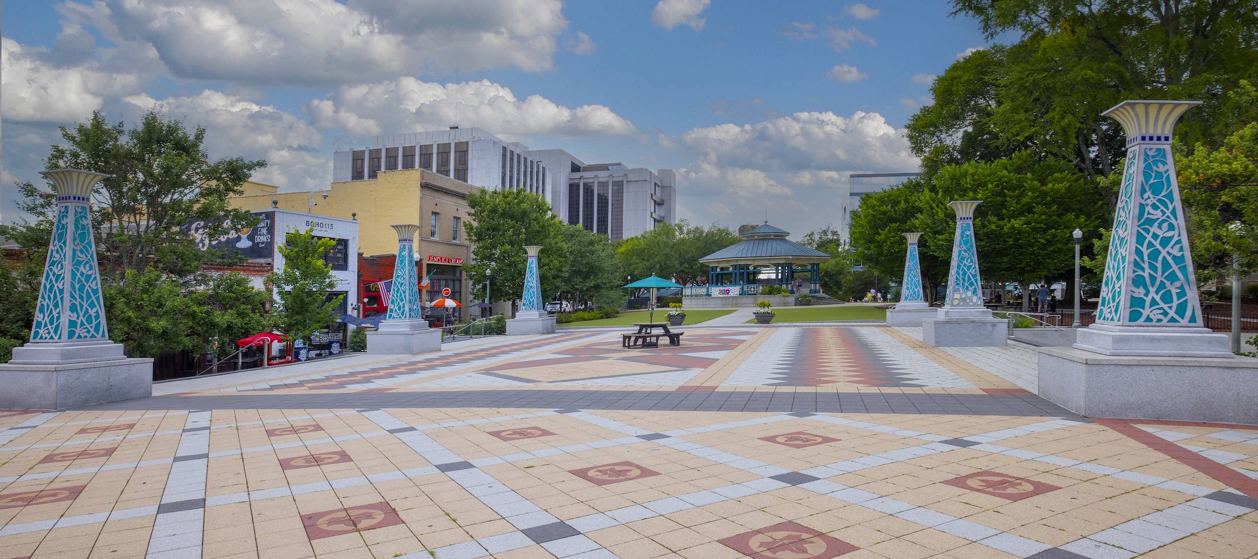 An outdoor plaza with decorative blue and white columns, a gazebo, trees, and a patterned tiled ground under a partly cloudy sky.