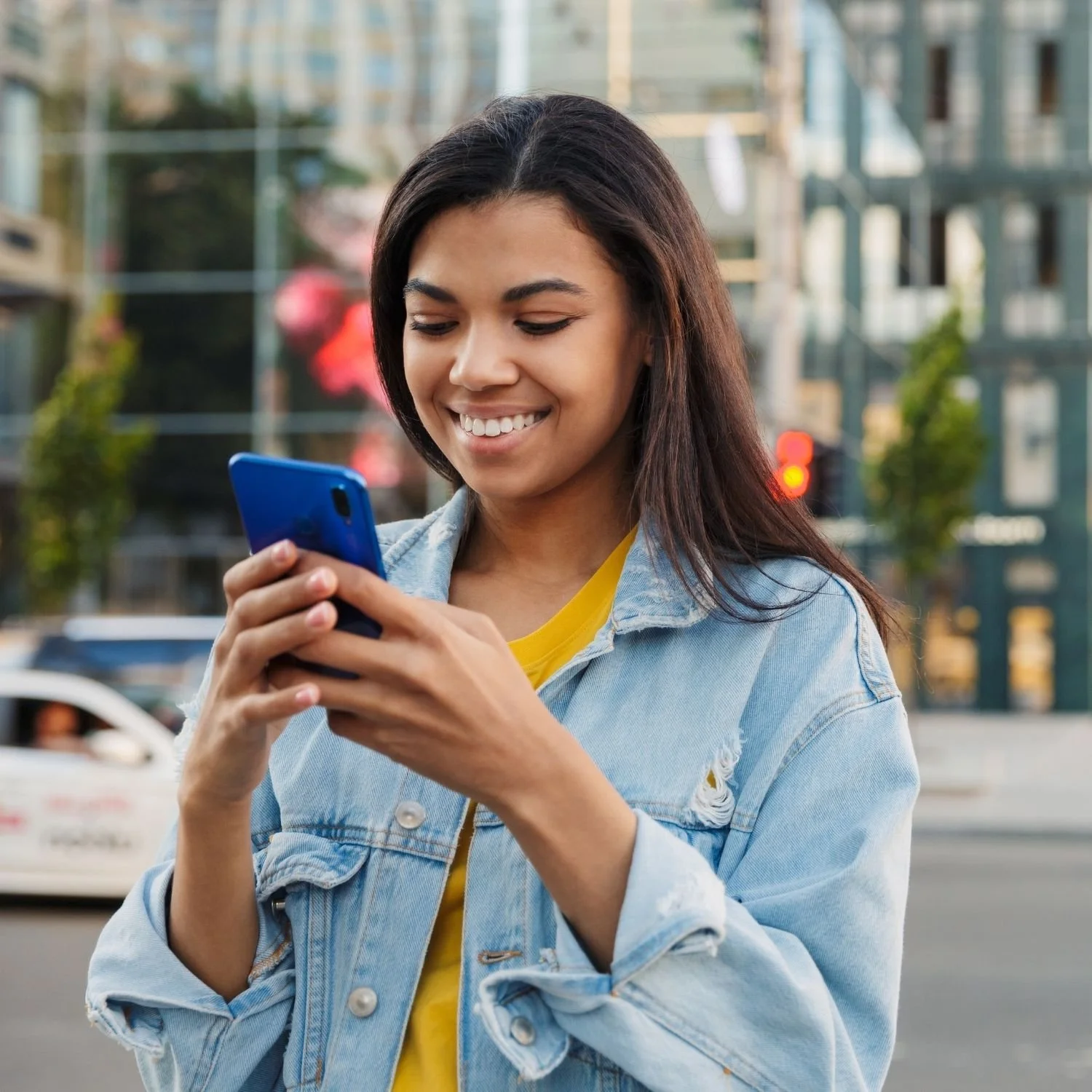 A young woman with dark hair smiling and looking at her smartphone while standing outdoors in an urban area.