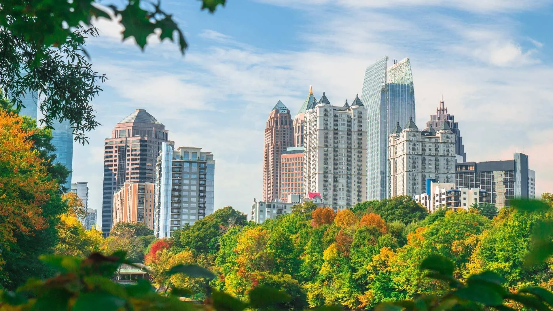 A city skyline with tall modern buildings viewed through lush green trees with autumn-colored foliage.