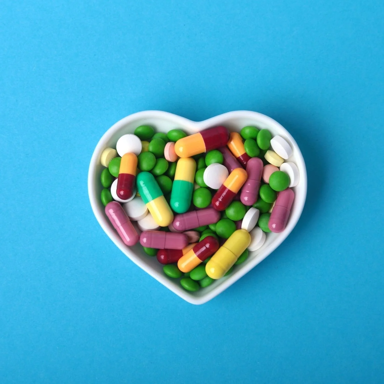 Heart-shaped white bowl filled with colorful capsules and tablets on a blue background.