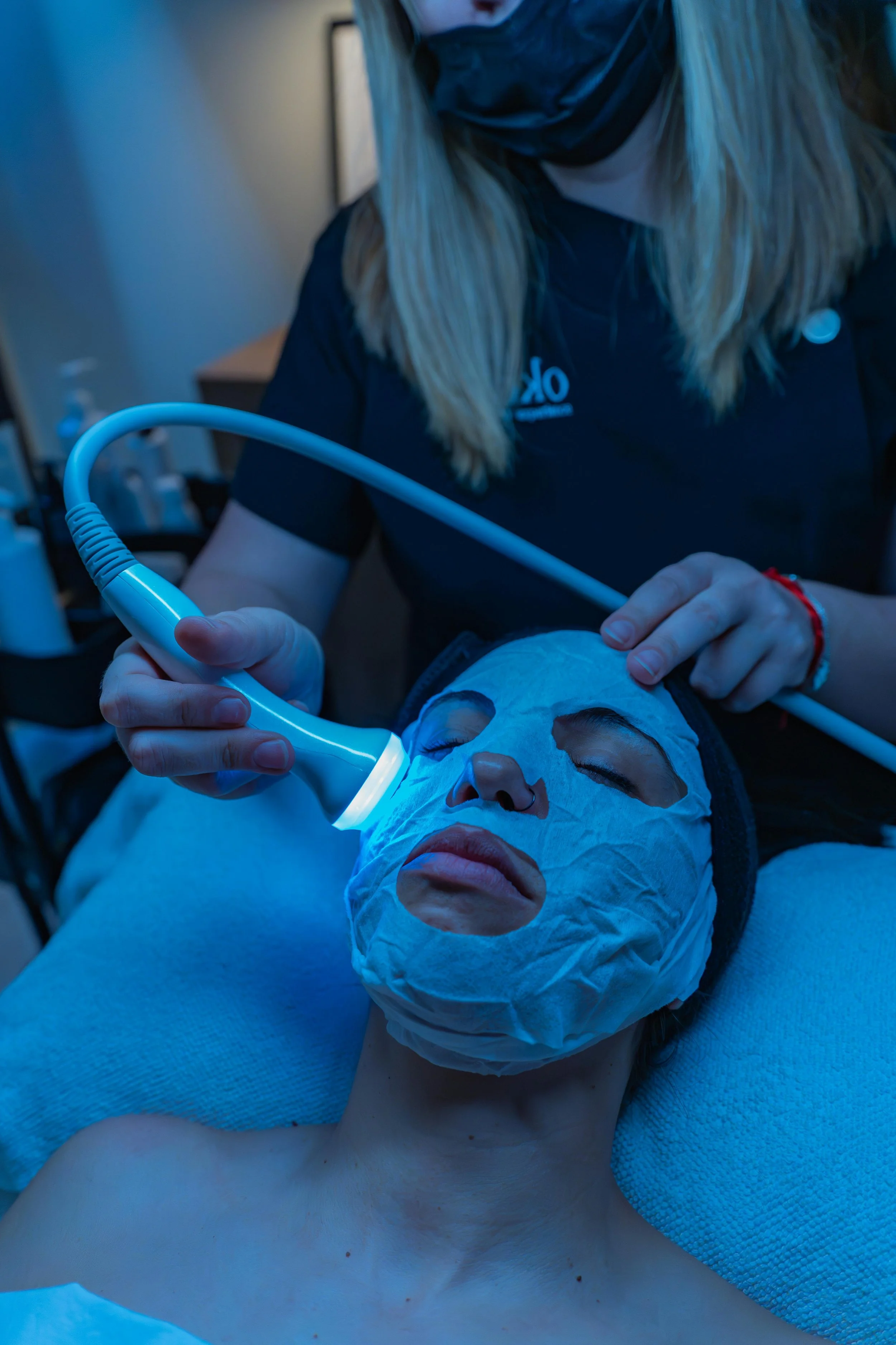 A woman is receiving a facial treatment with a handheld device emitting blue light. She is lying down with a face mask on, her eyes closed, while a practitioner holds the device near her face.