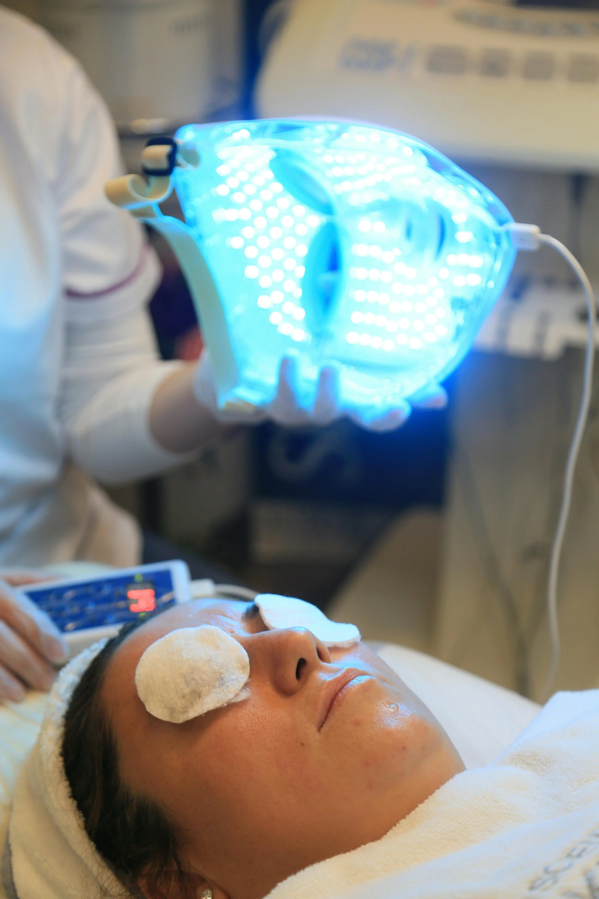 A person lying in a medical bed with eye pads, receiving a blue LED light therapy treatment while a healthcare professional holds a blue LED mask near their face.