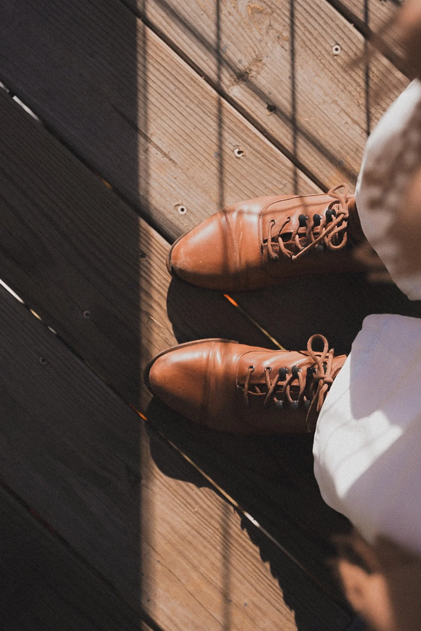 A person's feet wearing brown leather shoes and white pants, standing on a wooden floor with shadows cast across.