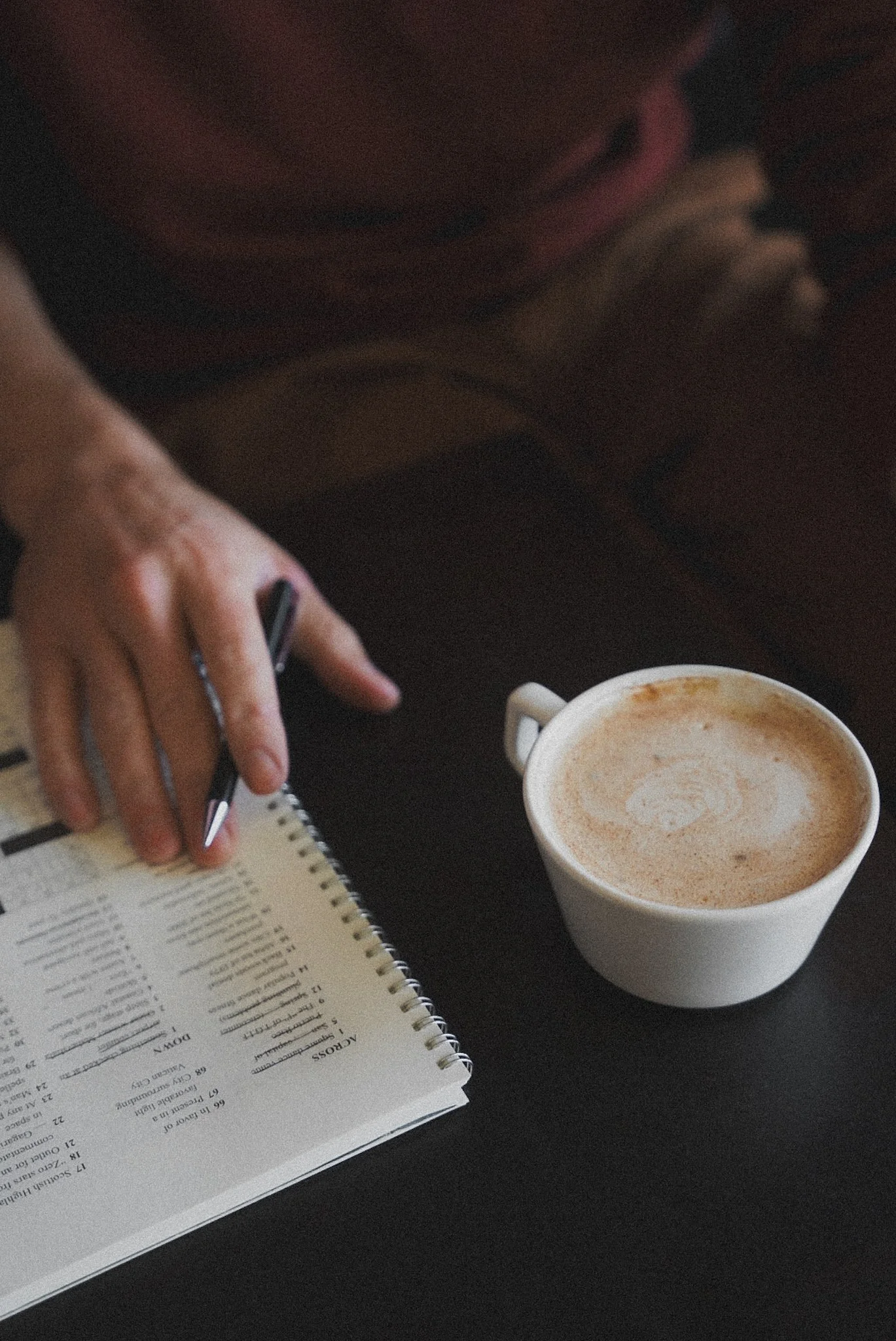 A person holding a pen and a notebook with a menu, next to a cup of coffee on a dark table.