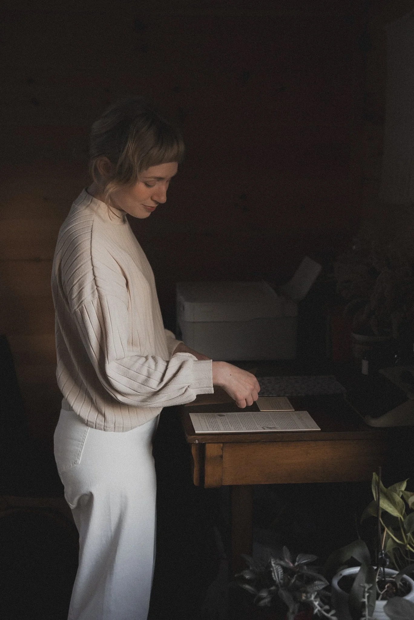 A woman with shoulder-length hair wearing a cream-colored, long-sleeved blouse and white pants, standing at a wooden desk in a dimly lit room, looking down at and reading from an open book.