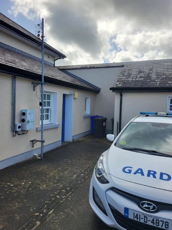 A parking area outside a house with a white Hyundai police car with blue markings and a Garda license plate in the foreground. The house has cream-colored walls, a blue door, and two windows, with some utility boxes and a black trash bin nearby. The sky is cloudy with patches of sunlight.