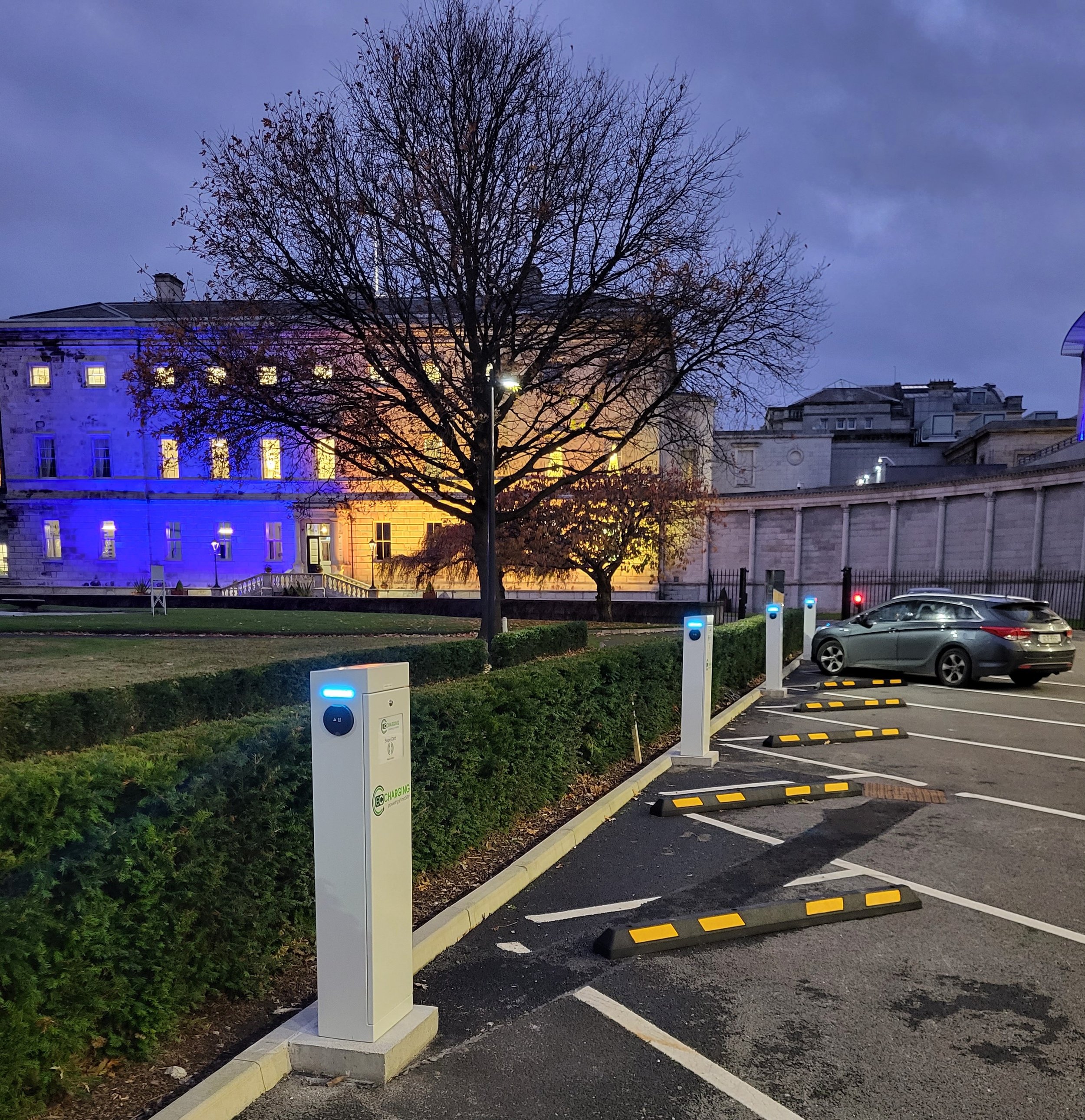 Electric vehicle charging station in a parking lot during evening with a tree and a historic building illuminated in the background.