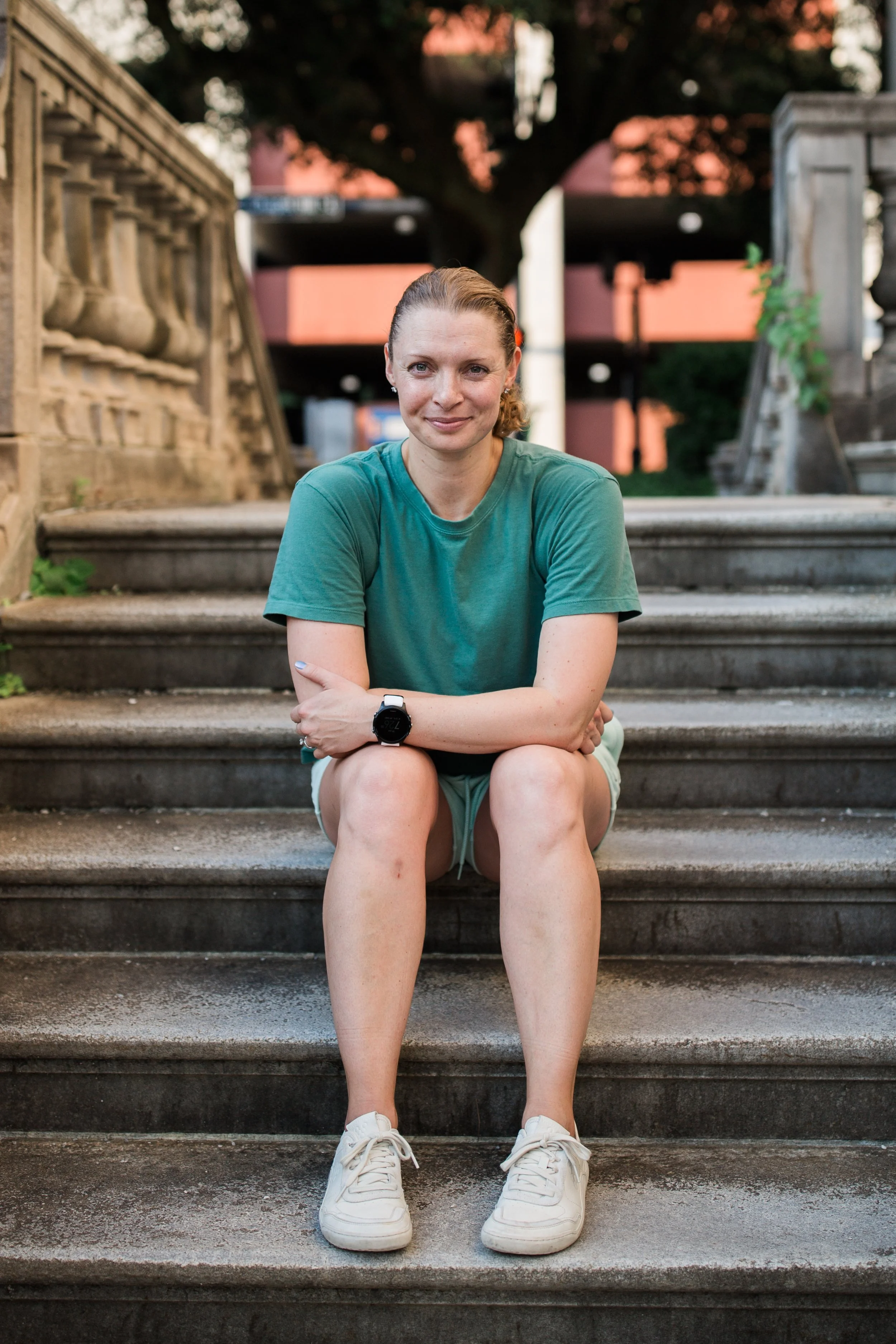 A woman with dirty blonde hair sitting on outdoor stone steps, wearing a teal t-shirt, shorts, white sneakers, and a black smartwatch, with a background of trees and modern buildings.