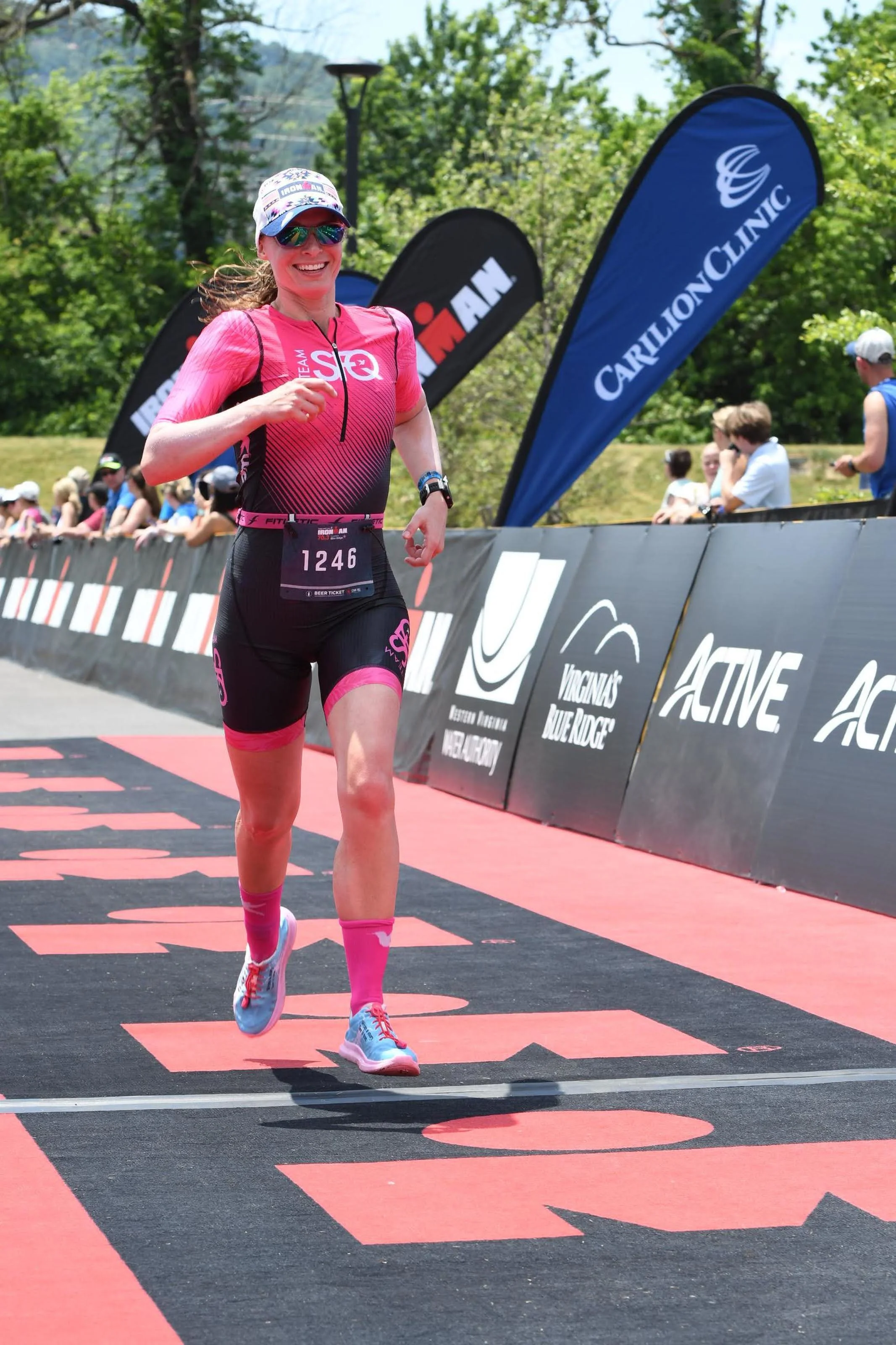 Female athlete running in a race, wearing a pink and black triathlon suit, sunglasses, and a hat, with a race bib numbered 1246. Spectators are visible behind the barriers, and promotional flags and banners are in the background.