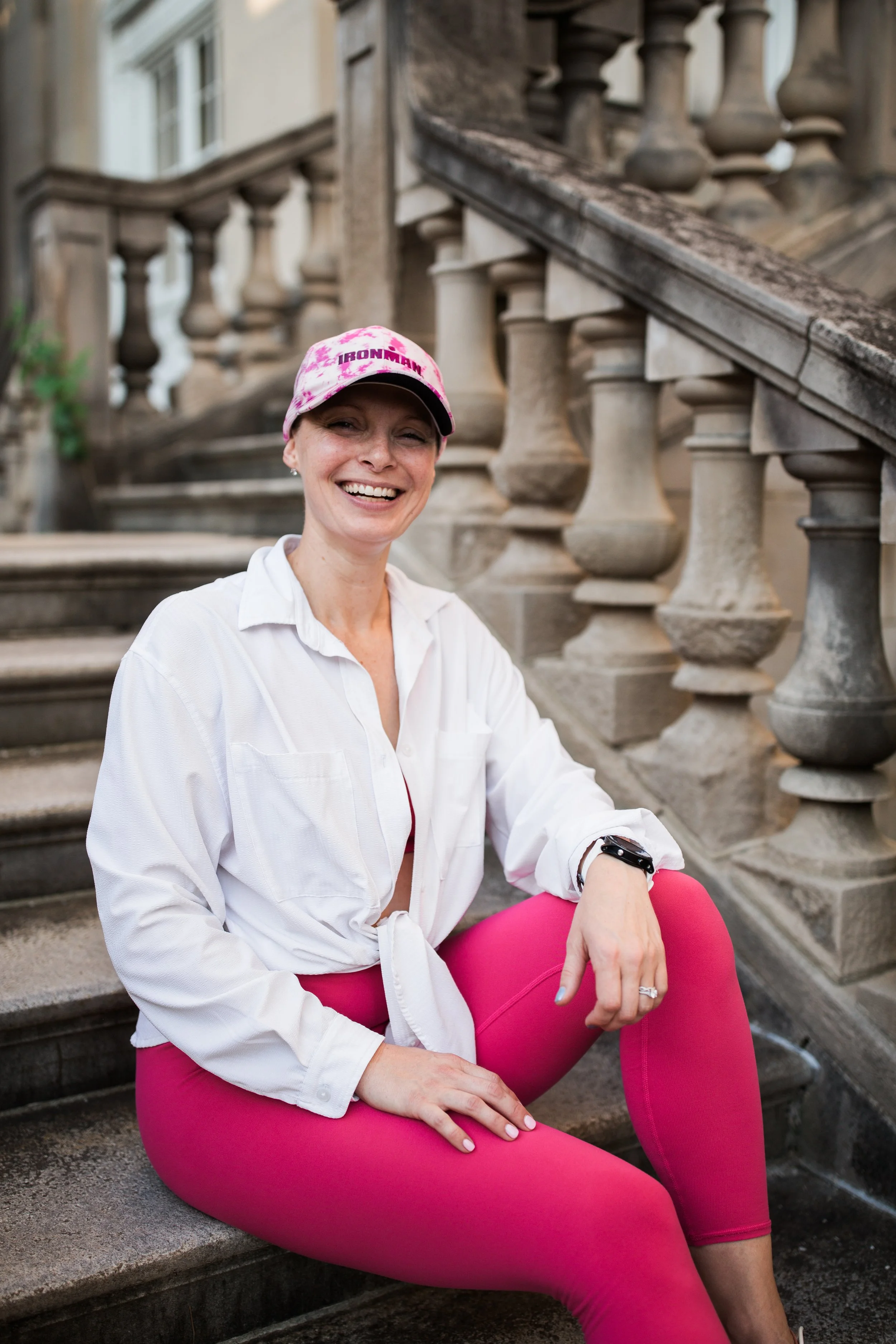 A woman sitting on stone steps outside a building, smiling, wearing pink leggings, a white shirt, a pink cap, and a fitness tracker.