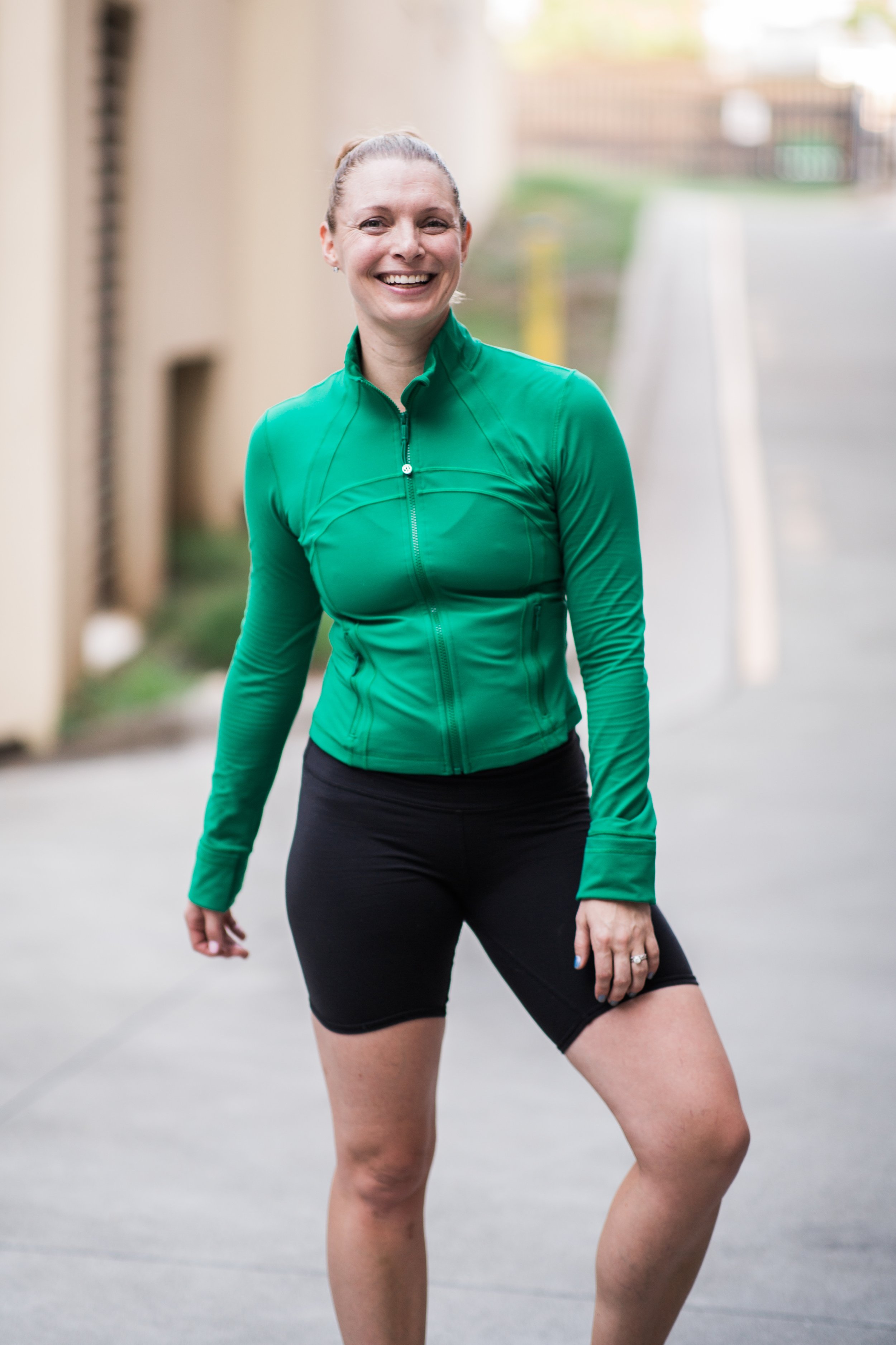 A smiling woman outdoors in athletic wear, wearing a green zip-up jacket and black shorts, standing on a sidewalk.