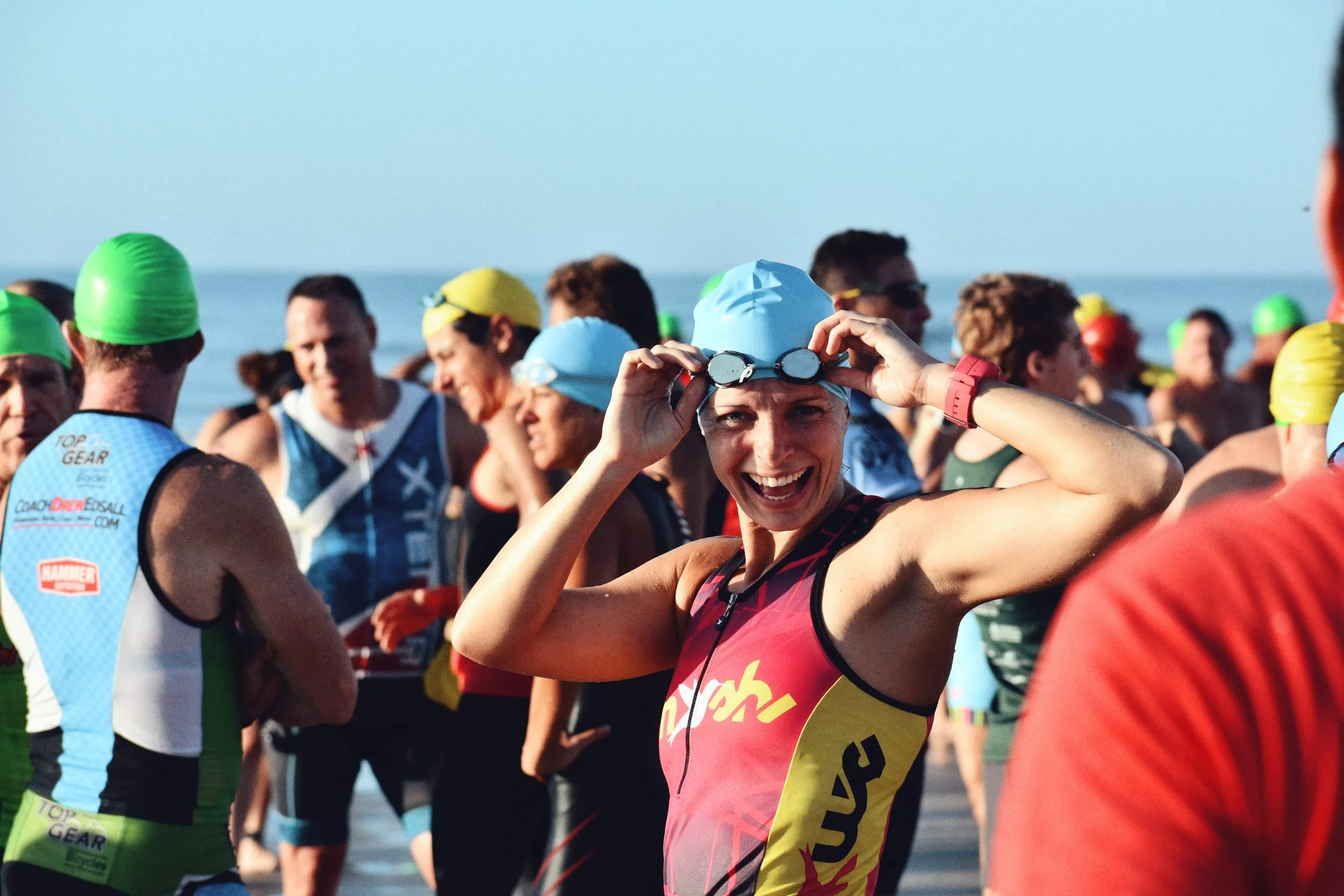 Smiling woman in a triathlon suit holding goggles, surrounded by other athletes at the beach.