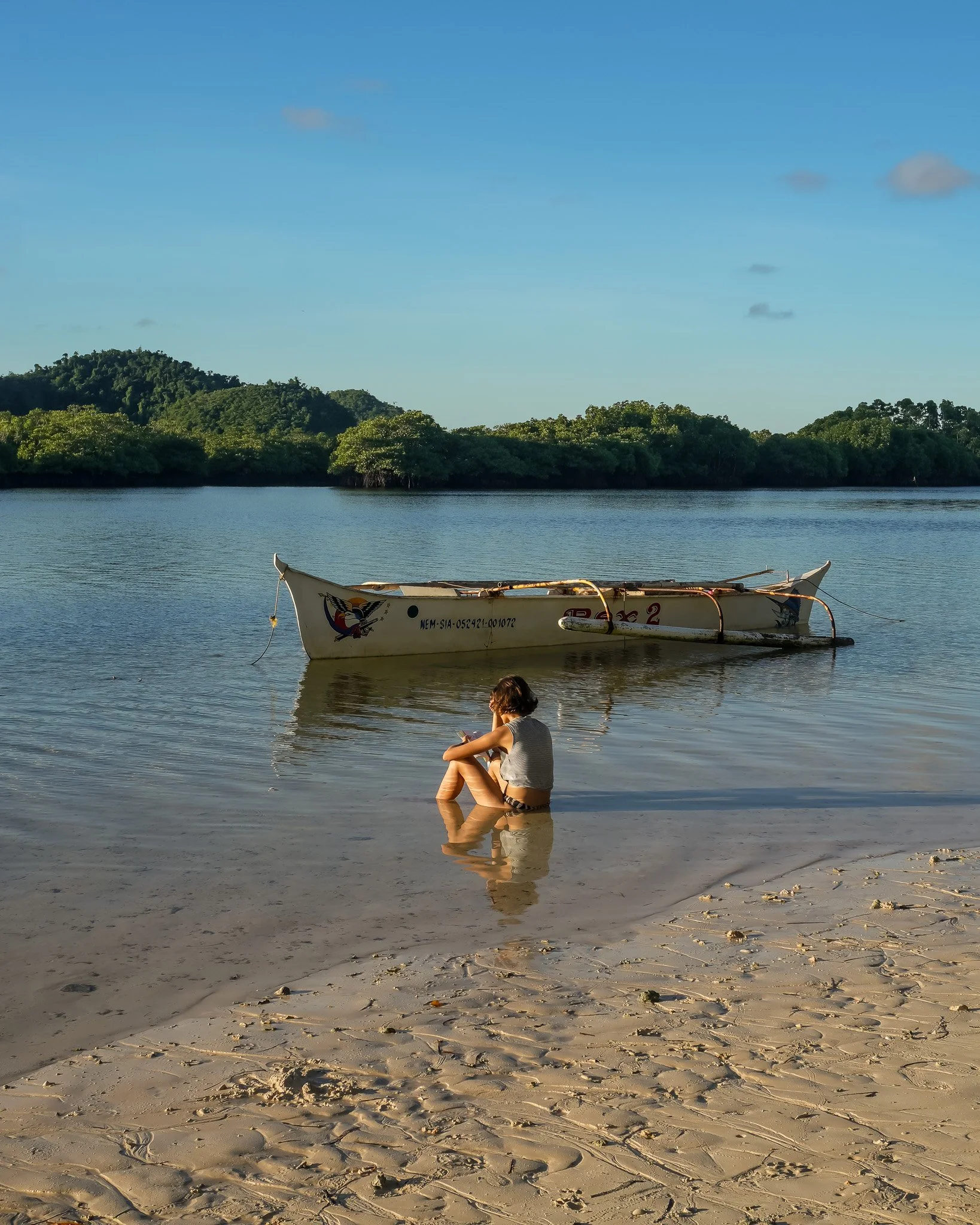 A woman sitting at the edge of the water on a sandy beach, looking at a boat floating on calm water, with green trees and hills in the background under a clear blue sky.