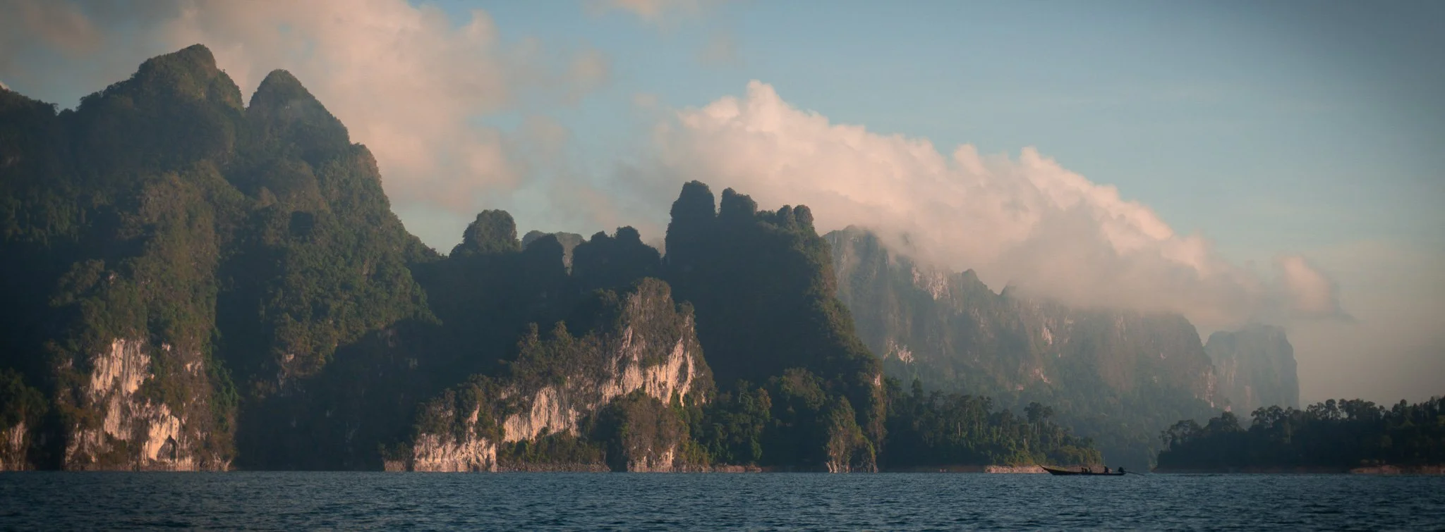 Lush green mountains rise sharply from a body of water with a boat floating on it, under a partly cloudy sky.