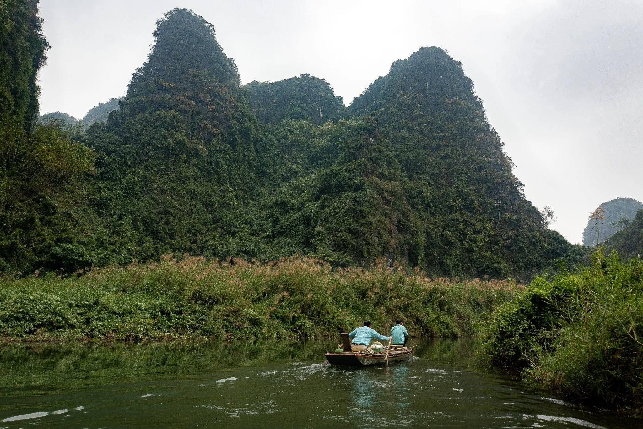 Two men in a small boat on a river, surrounded by green mountains and lush vegetation.