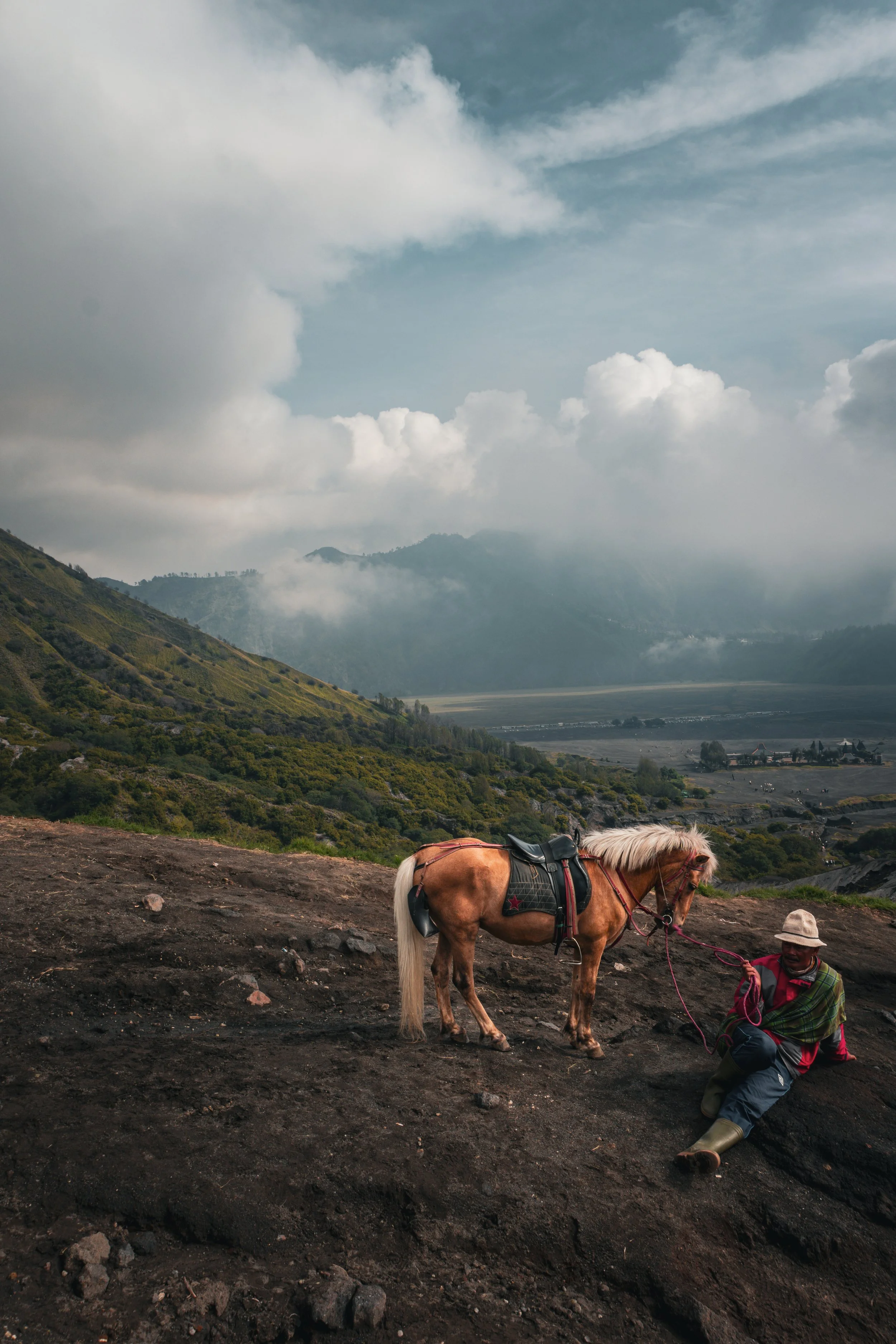 A man with a hat and poncho sitting on the ground holding a lead connected to a small horse with a saddle, on a landscape with mountainous terrain and cloudy sky.