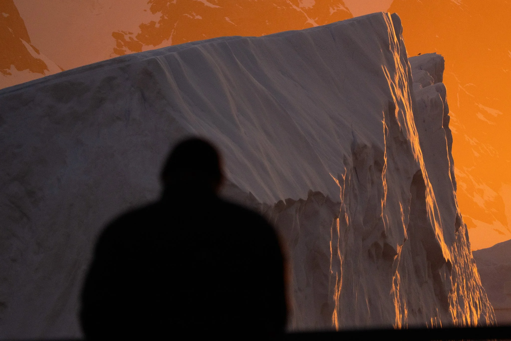 Silhouette of a person looking at a large icy glacier with a sunset sky in the background.