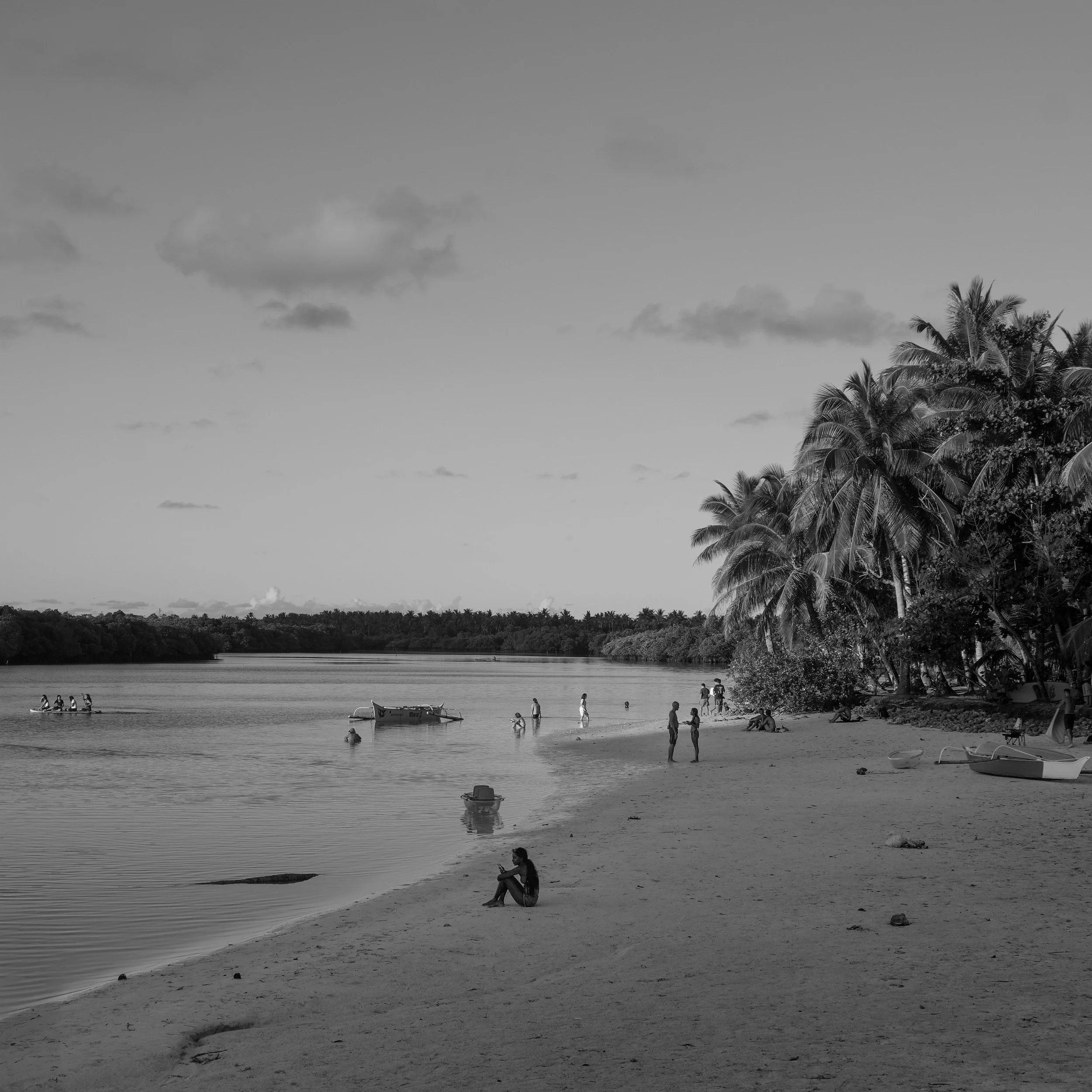 Black and white photo of a tropical beach with palm trees, people enjoying the shore, boats in the water, and a child sitting on the sand.