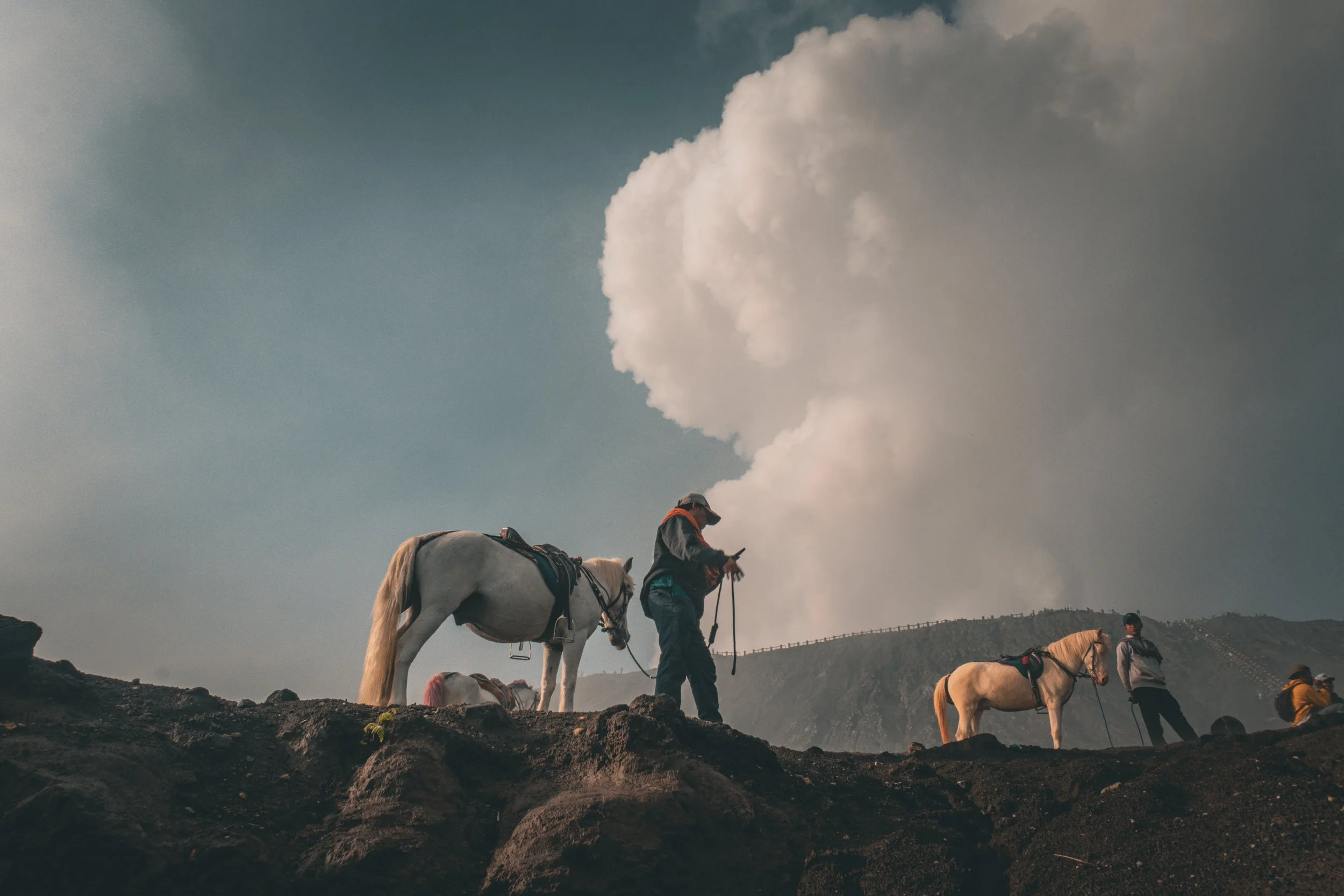 People and horses on a rocky terrain with a large cloud of smoke or ash in the background.