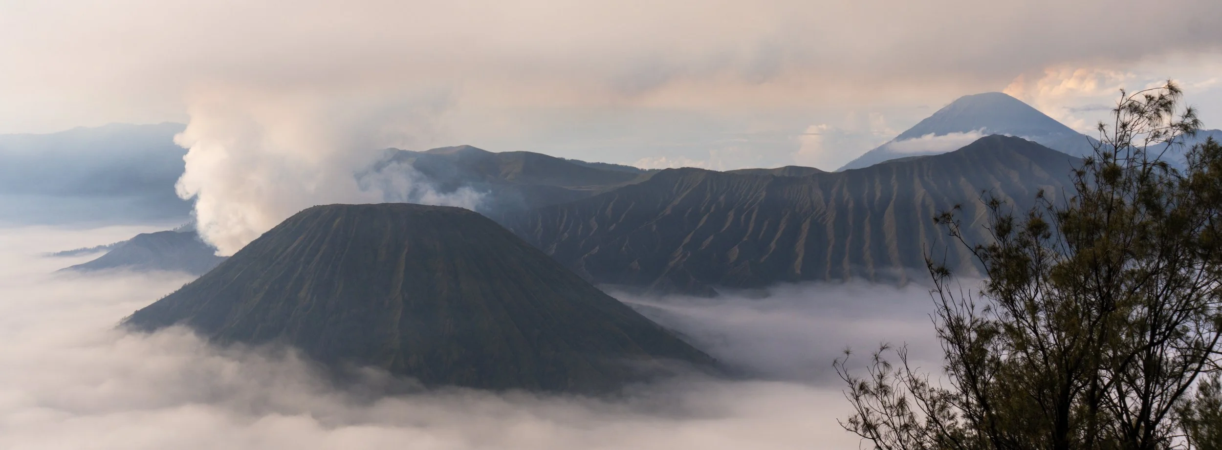 Mt. Bromo emitting smoke and steam with clouds surrounding them, including a tall mountain in the background.