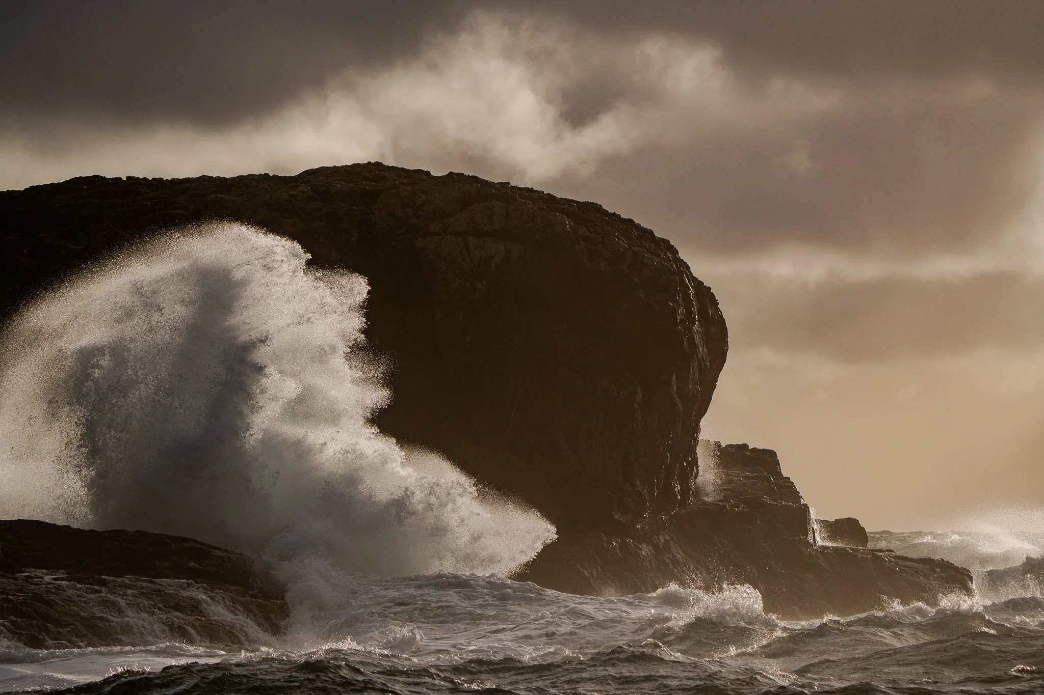 Ocean waves crashing against large rocks under dark, cloudy sky during stormy weather.