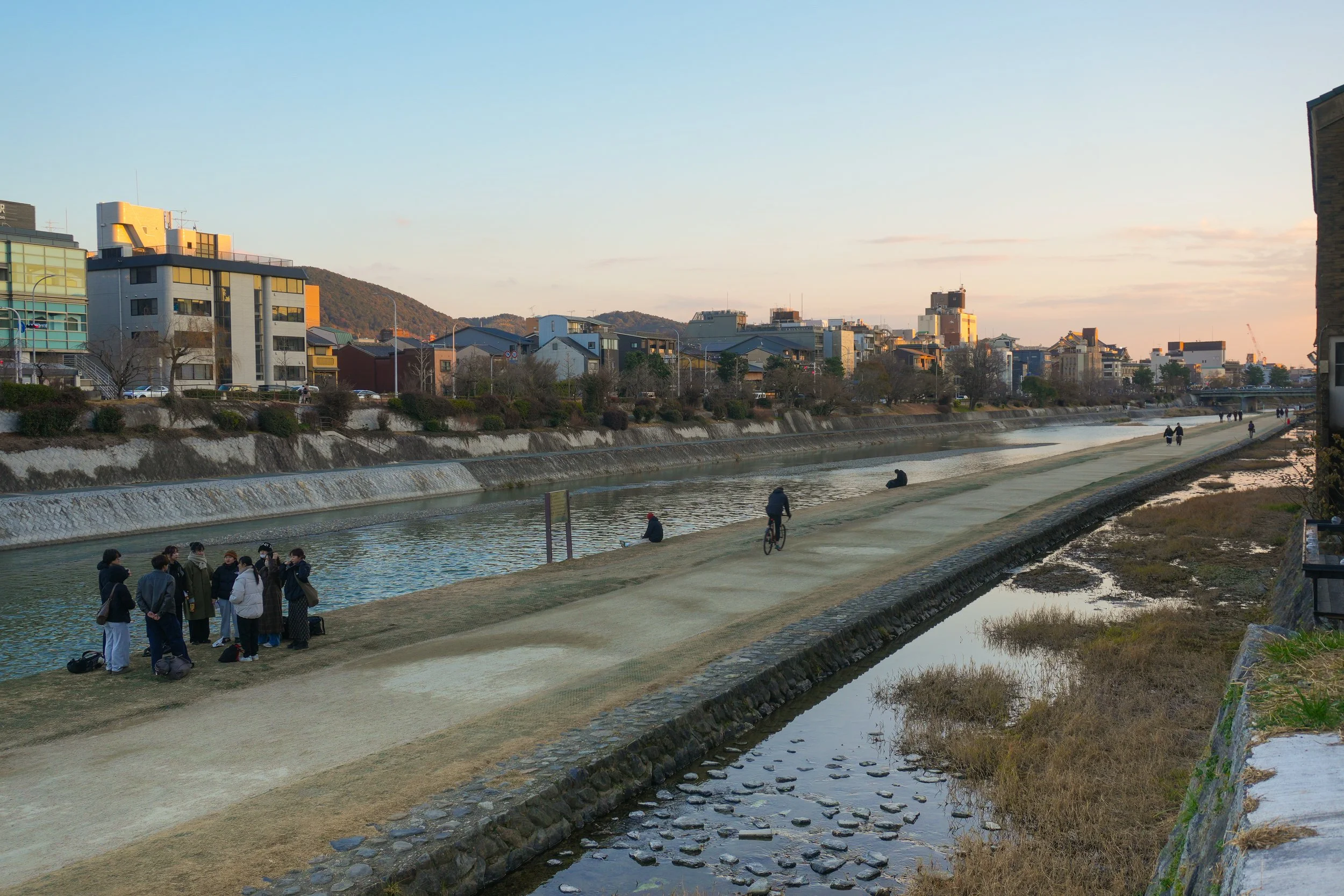 People gather and walk along a riverbank in an urban area during sunset, some sitting, some walking, and one person riding a bike, with buildings and a hill in the background.
