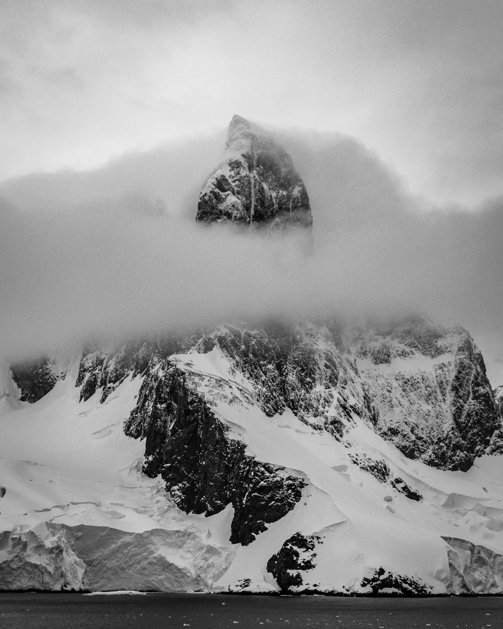 A black and white photograph of a snow-covered mountain with a pointed summit, partially obscured by clouds or fog, and a body of water at the base.