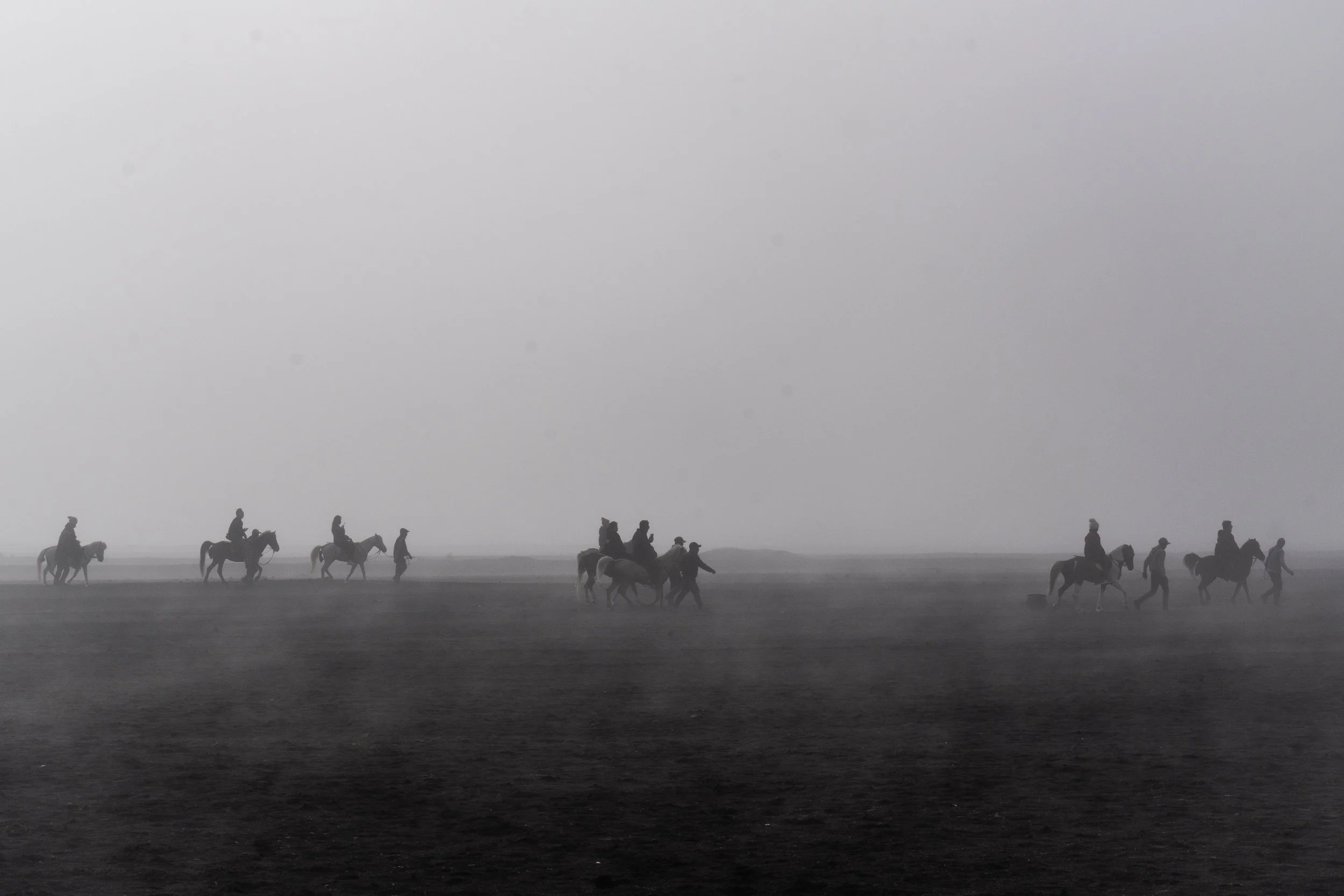 A line of people riding horses across a foggy landscape in black and white.