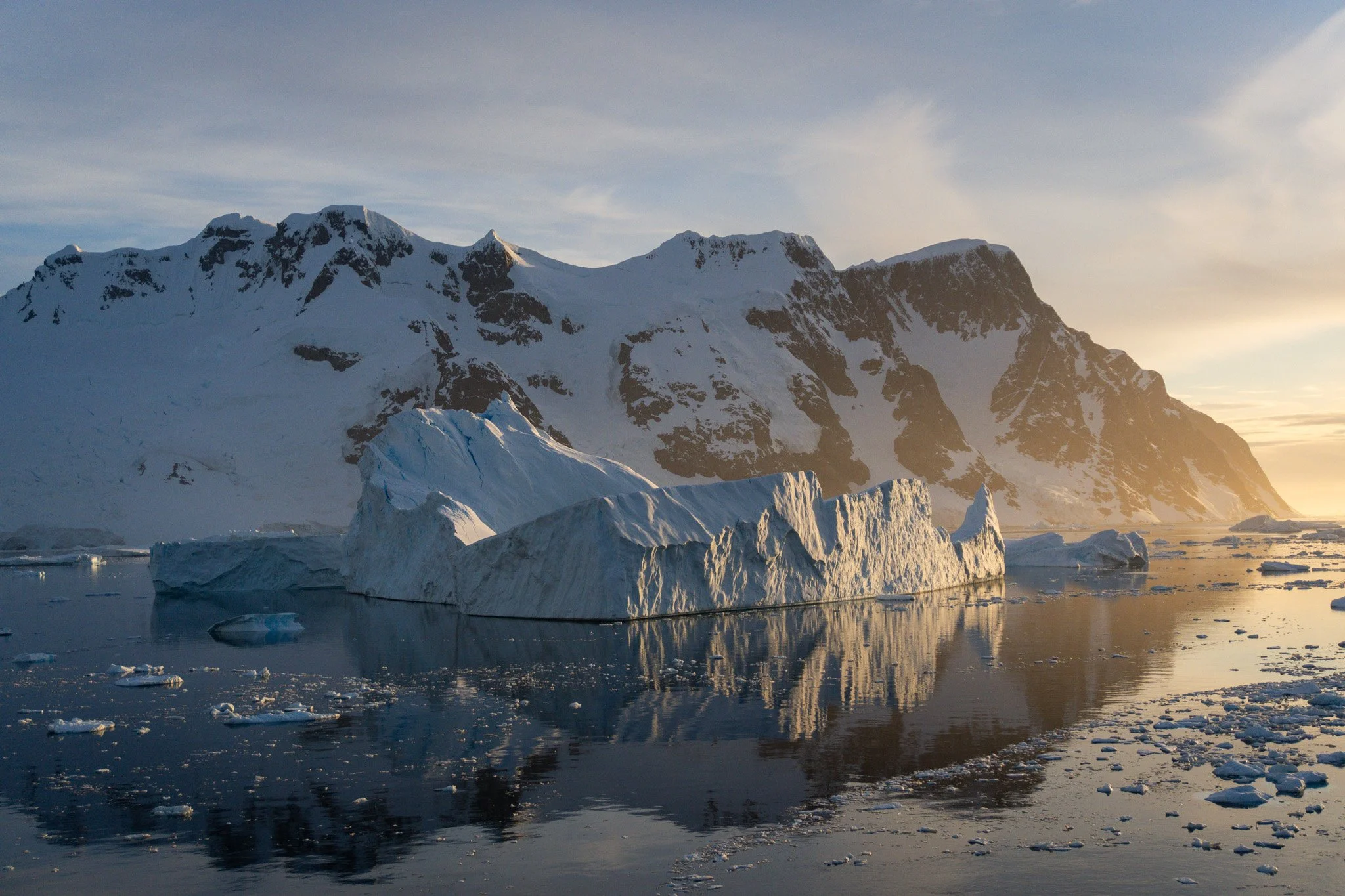 A landscape of snow-covered mountains with a large iceberg floating in the water in the foreground, during sunset or sunrise with a golden sky.