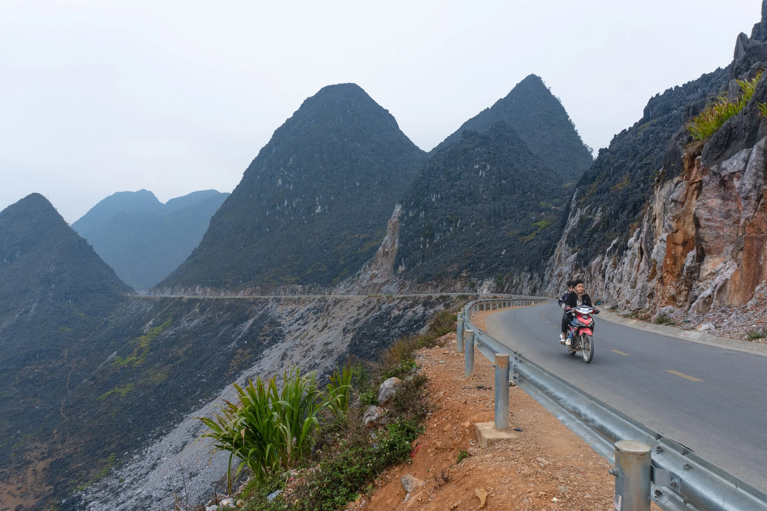 A winding mountain road with a person riding a motorcycle along the guardrail, surrounded by tall, rugged mountains under an overcast sky.