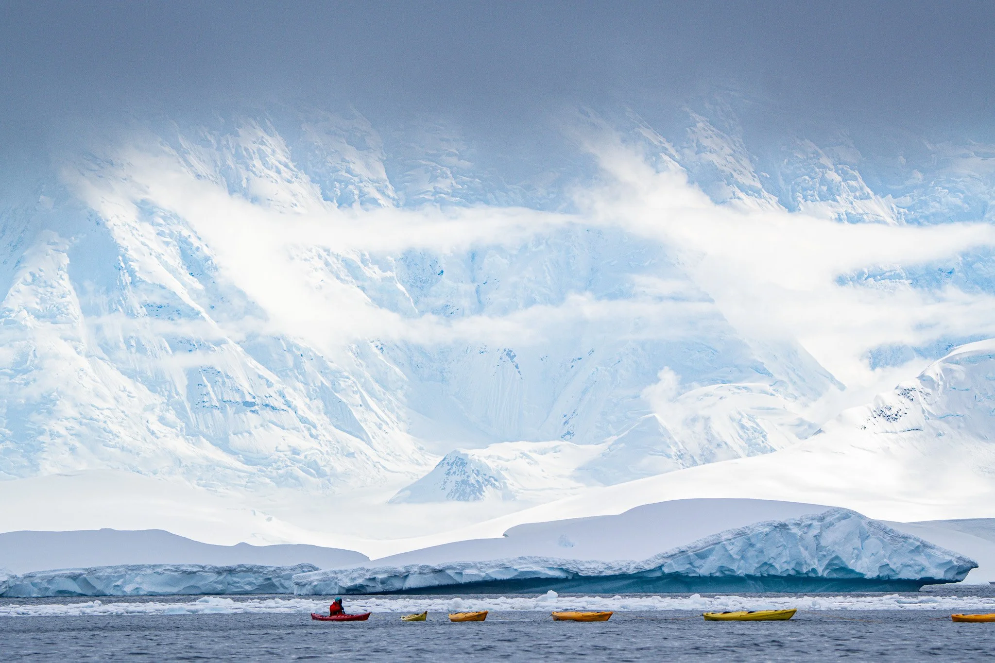 A person in a red jacket on a kayak near yellow kayaks in icy water, with a large ice formation and snow-covered mountains and glaciers in the background.