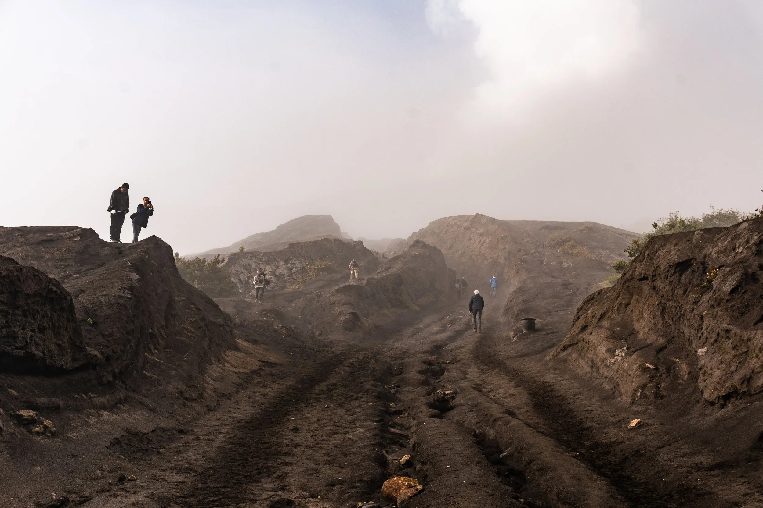Several people exploring a volcanic or rocky terrain with dark soil, with some taking photos and others walking along dirt paths. The sky is cloudy and foggy, obscuring the background mountains.