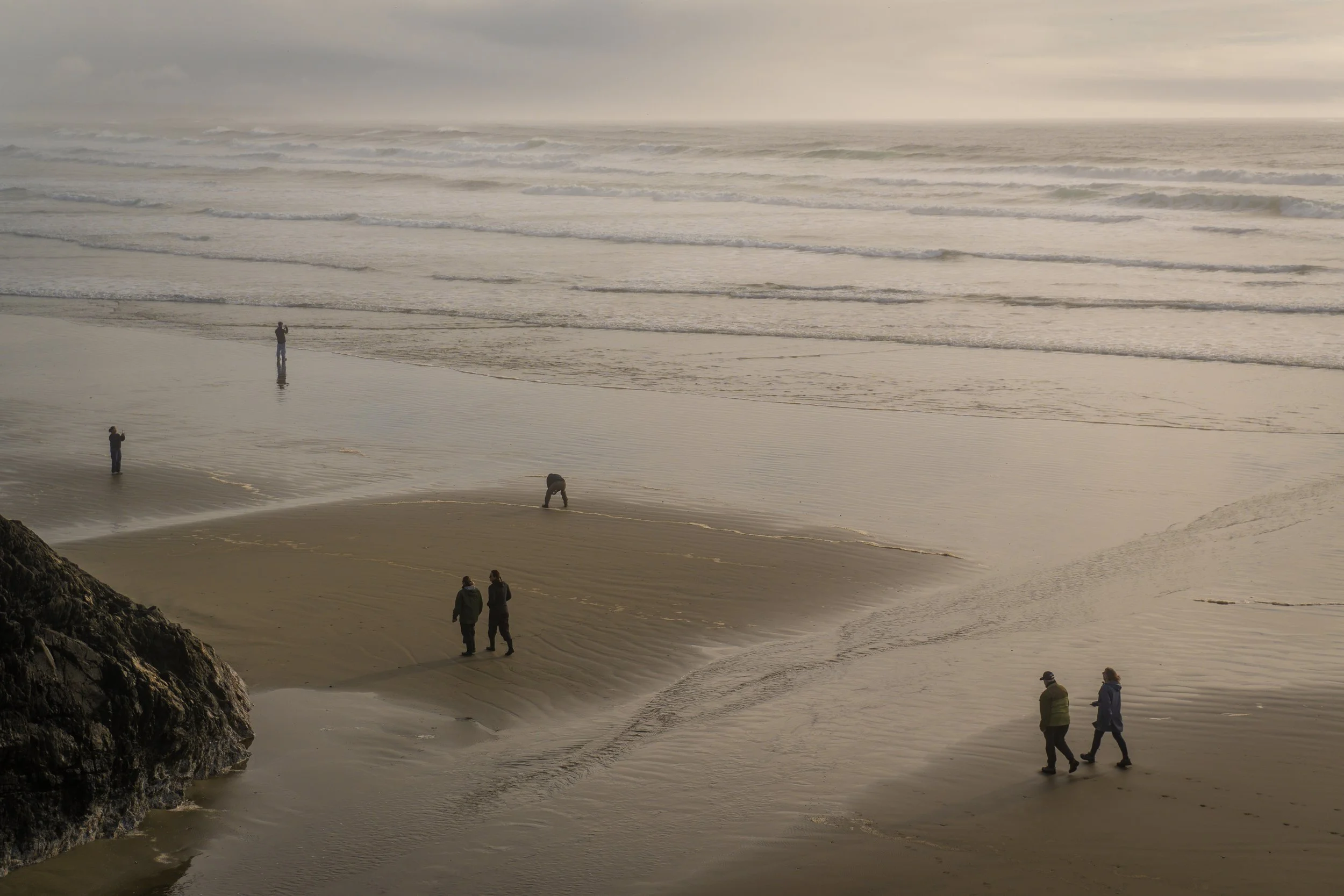 People walking and standing on a sandy beach with waves in the ocean and a cloudy sky.